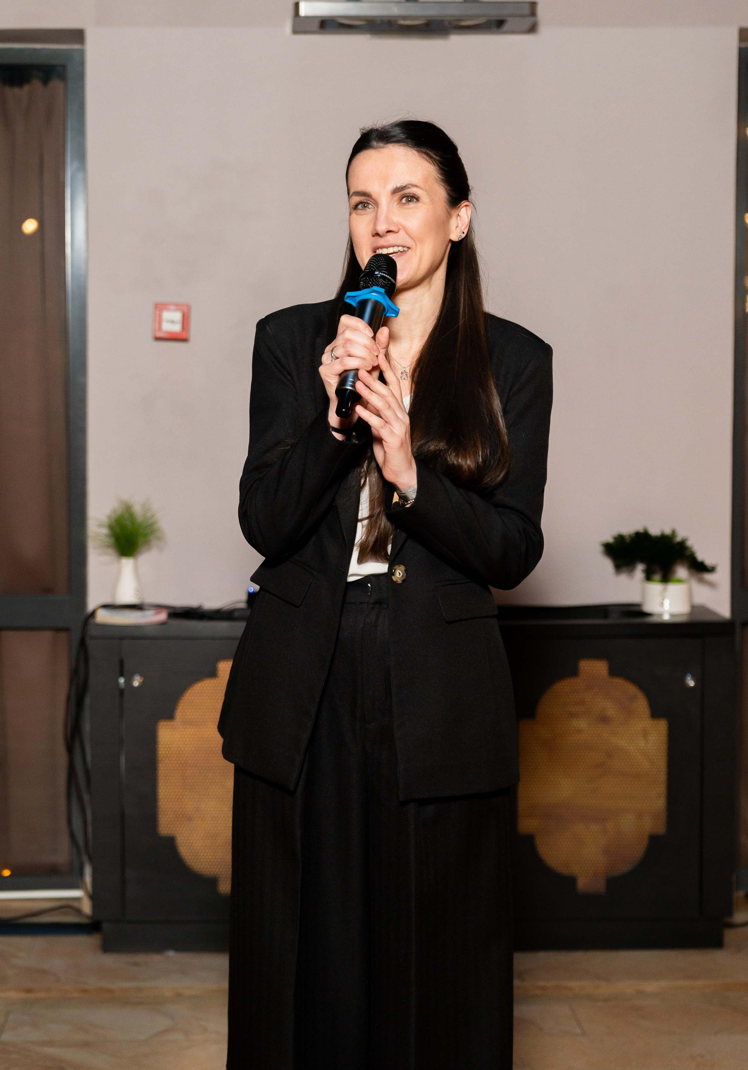 Женщина в чёрном костюме выступает с микрофоном на мероприятии Radisson Sonya. Фотография Евгения Мальцева.