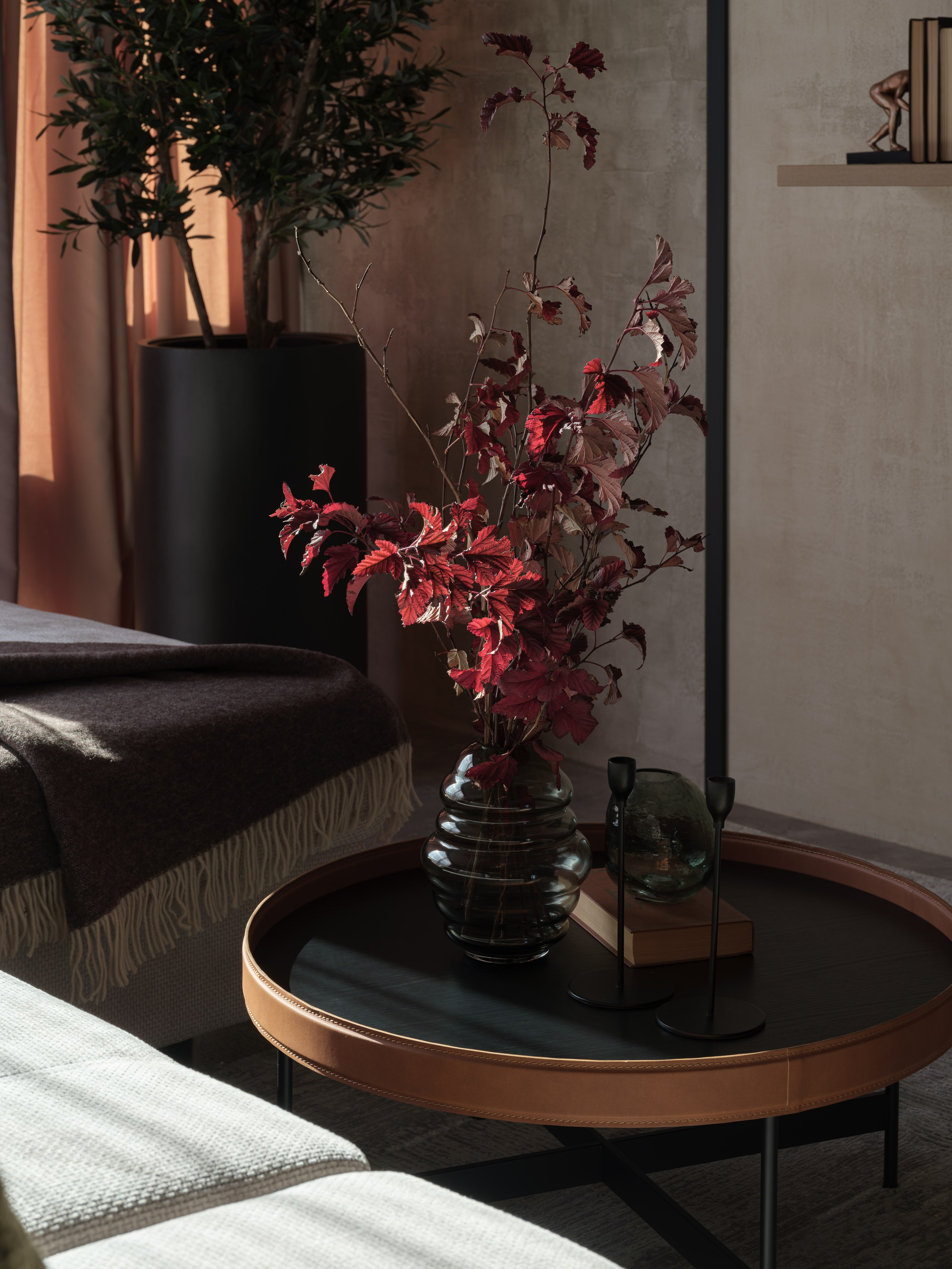Декоративная композиция в гостиной — стеклянная ваза с алыми ветвями на стильном журнальном столике с кожаной окантовкой. Тёплый свет и мягкий текстиль создают атмосферу уюта. Интерьер оформлен дизайнером Алиной Дёровой, стилист Наталья Якушева, фото Сергей Денисенко.