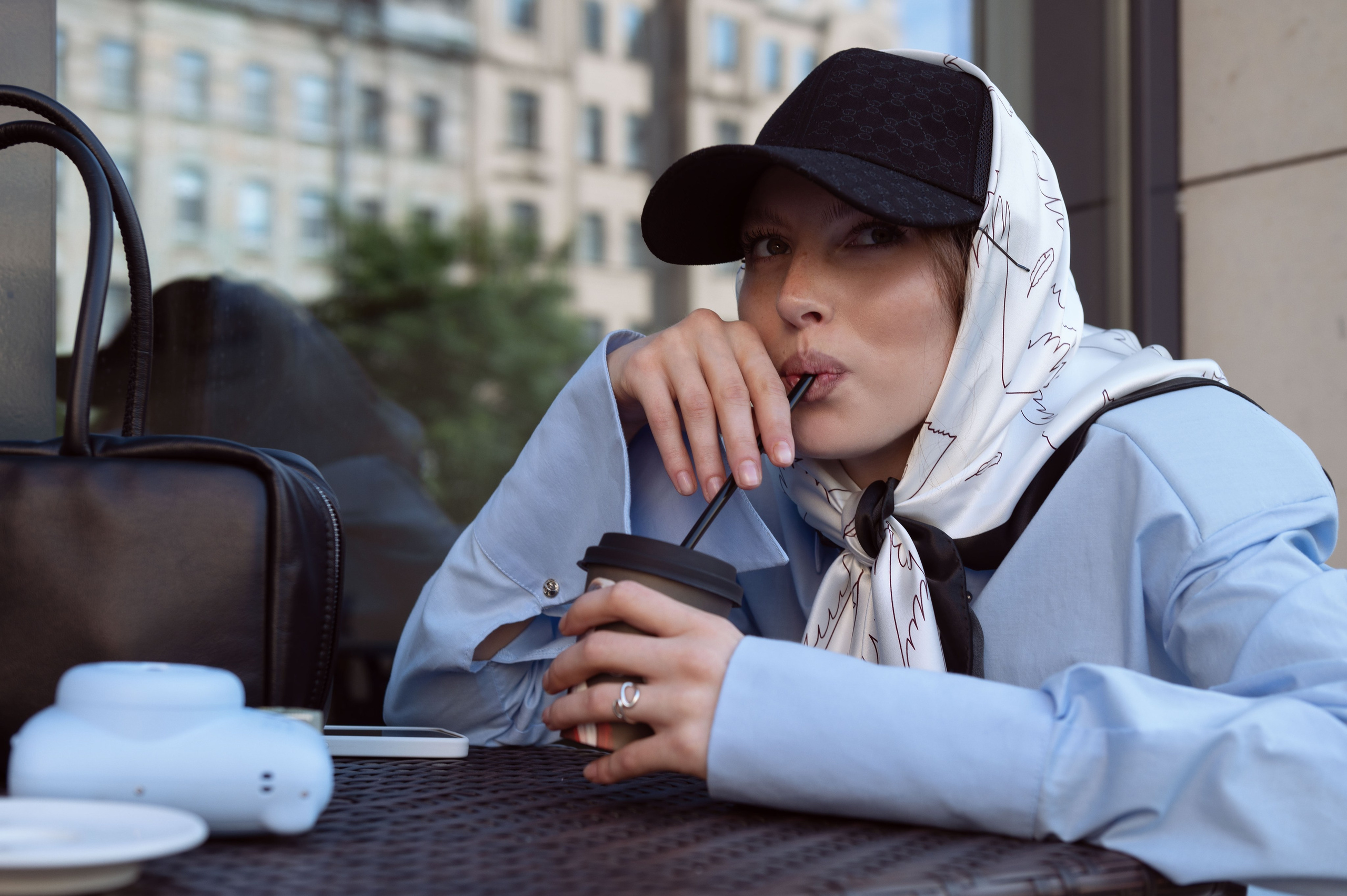 Красивая девушка в голубой рубашке и белых брюках и шелковом дизайнерском платке, повязанном поверх кепки сидит за столиком в уличном кафе и пьет напиток