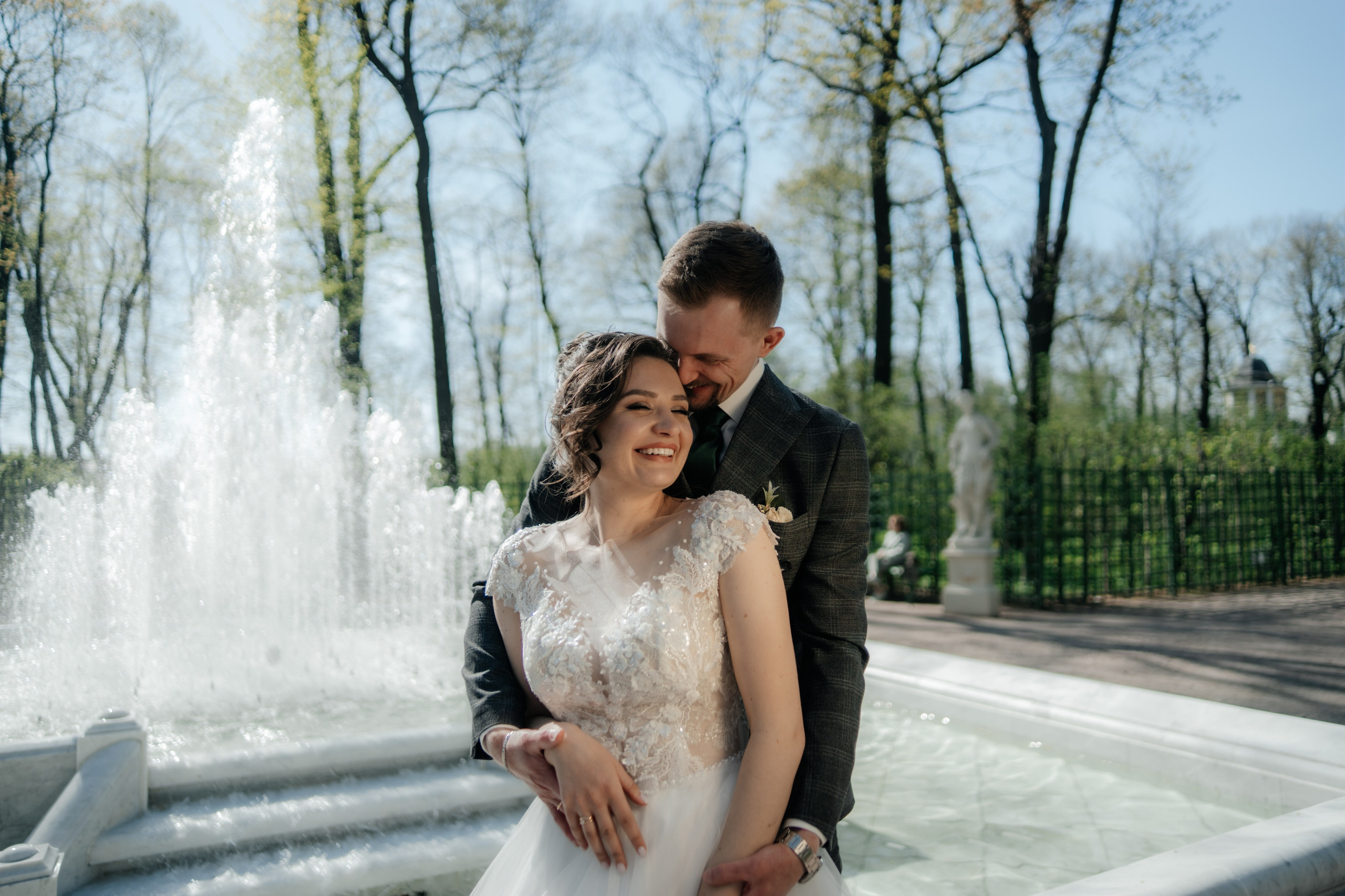 жених и невеста молодожены обнимаются около фонтана в летнем саду