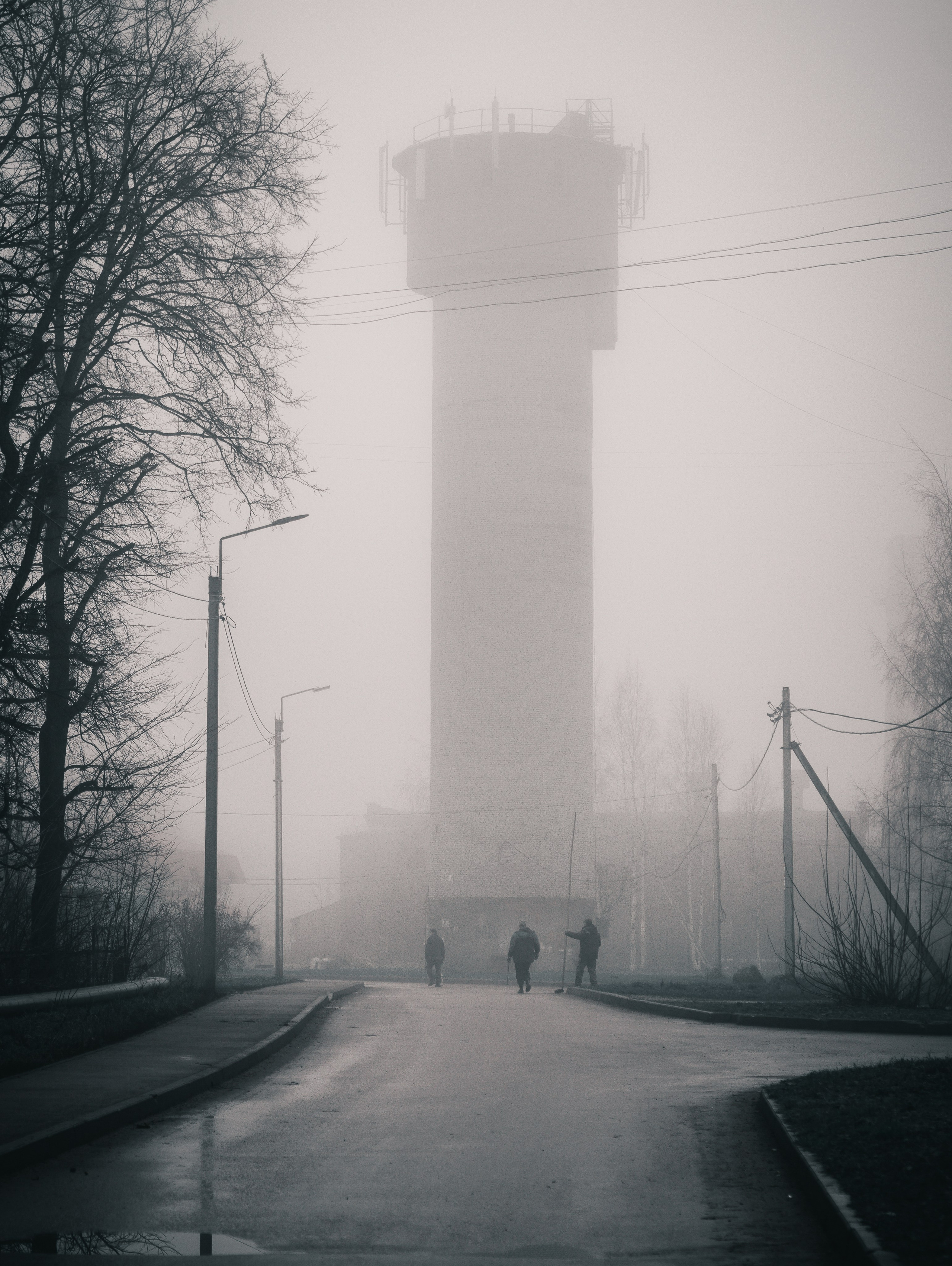 Туманное утро в промышленной зоне Санкт-Петербурга, водонапорная башня в густом тумане, трое рабочих с метлами идут по дороге, атмосферная уличная фотография, фотограф Евгений Мальцев (Evrednix).