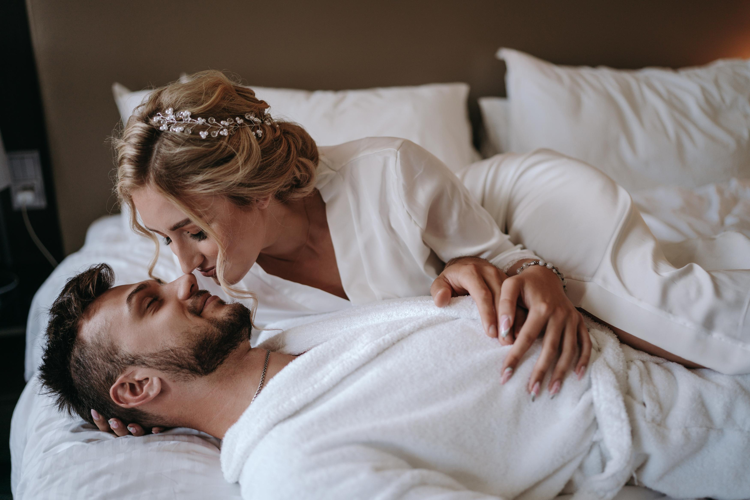 Невеста целует жениха который лежит на кровати в отеле, оба в халатах, классная идея для сборов жениха и невесты в номере отеля Шератон в Уфе