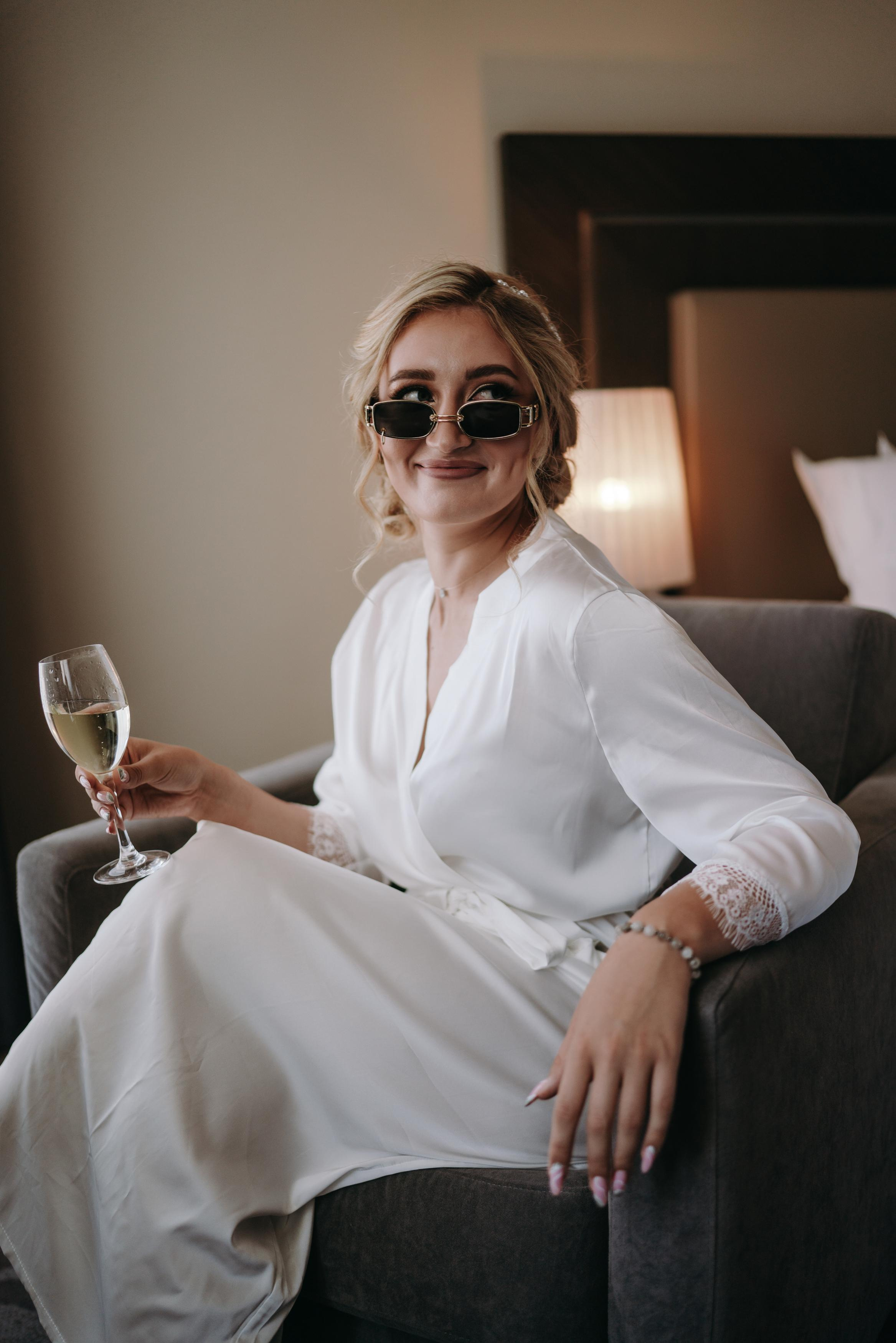 Красивая невеста сидит в халате и очках в номере отеля Шератон и пьет шампанское