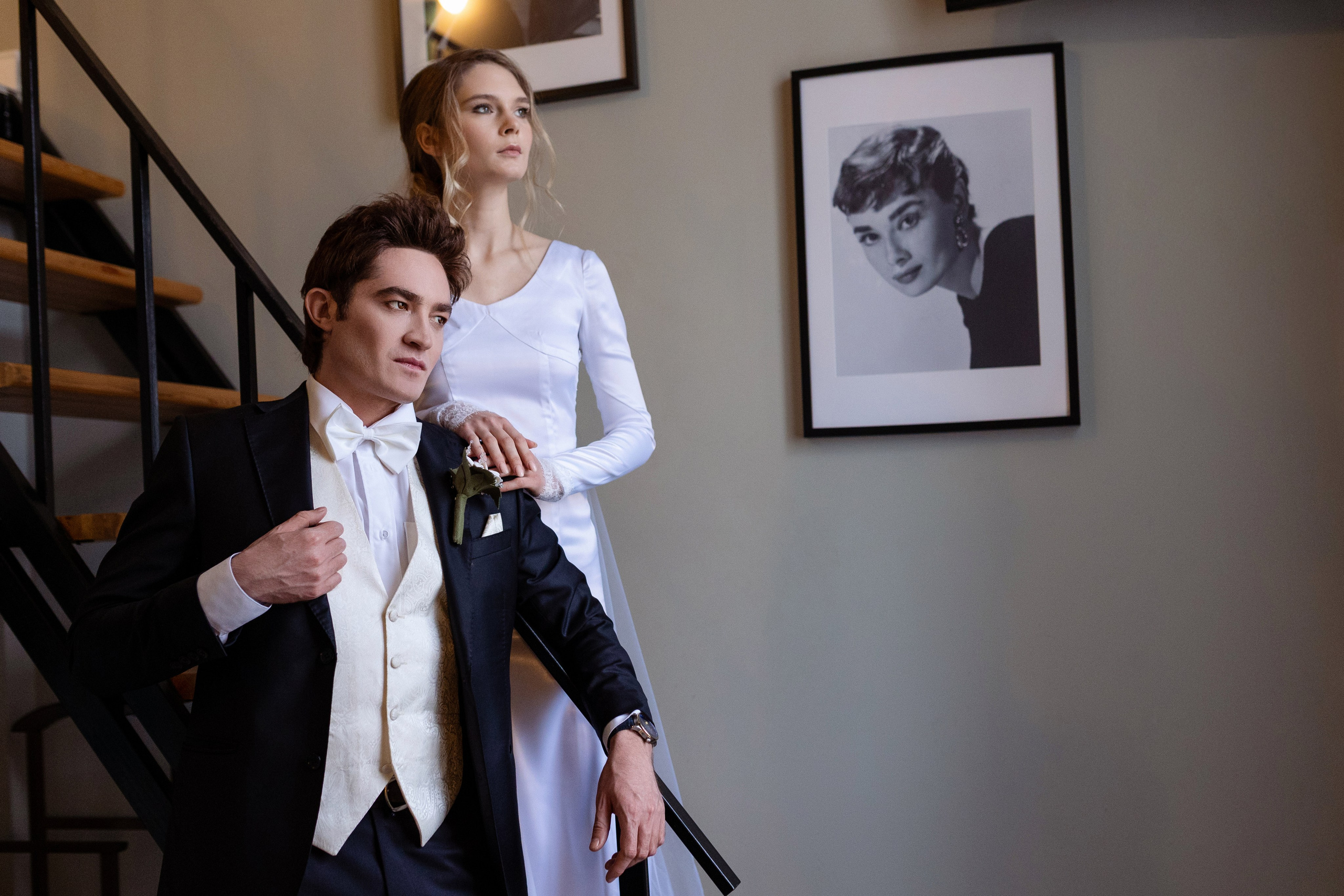 Невеста стоит на лестнице  сзади жениха. на  стене висит фотография.