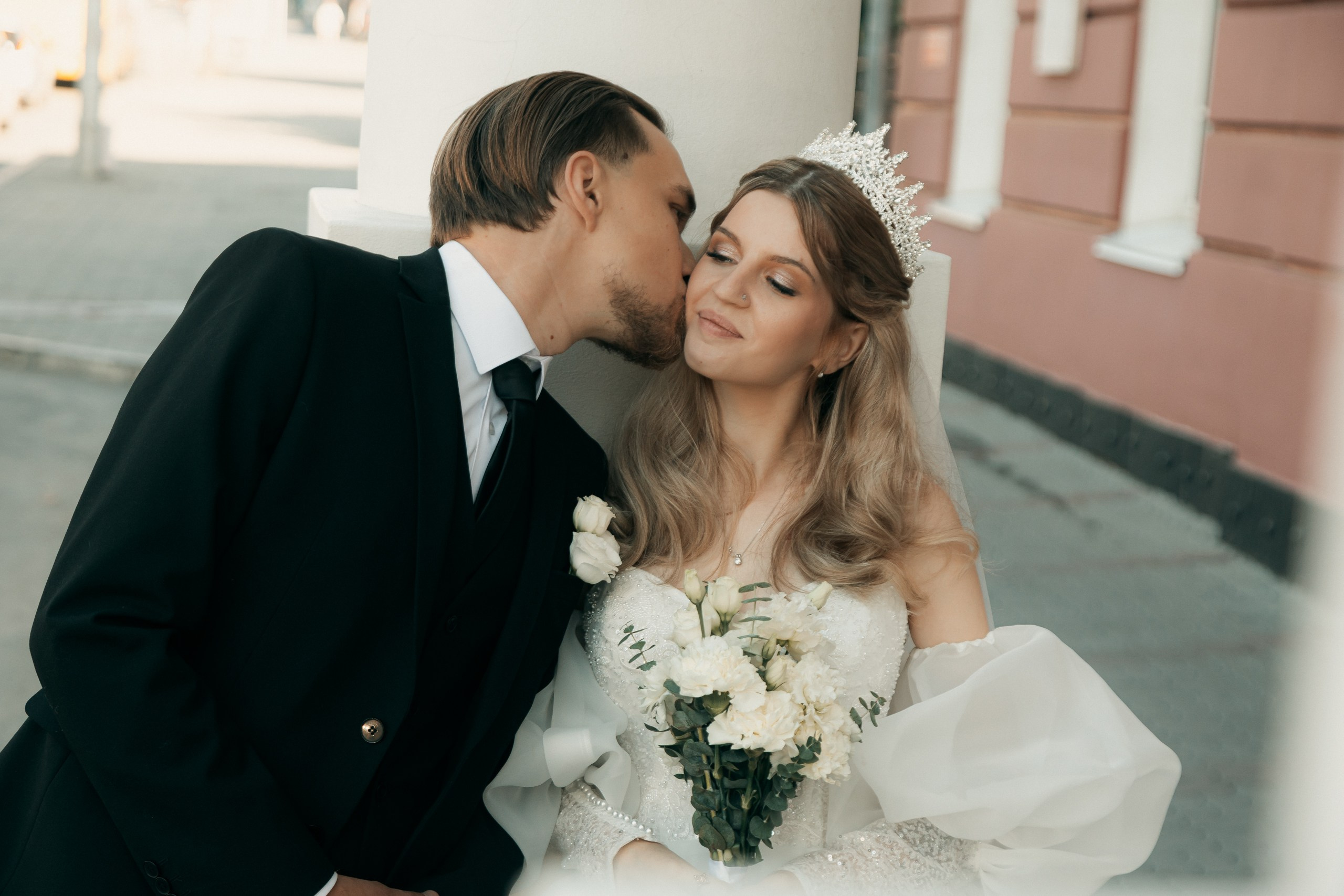 Жених целует невесту в щеку. Невеста улыбается.