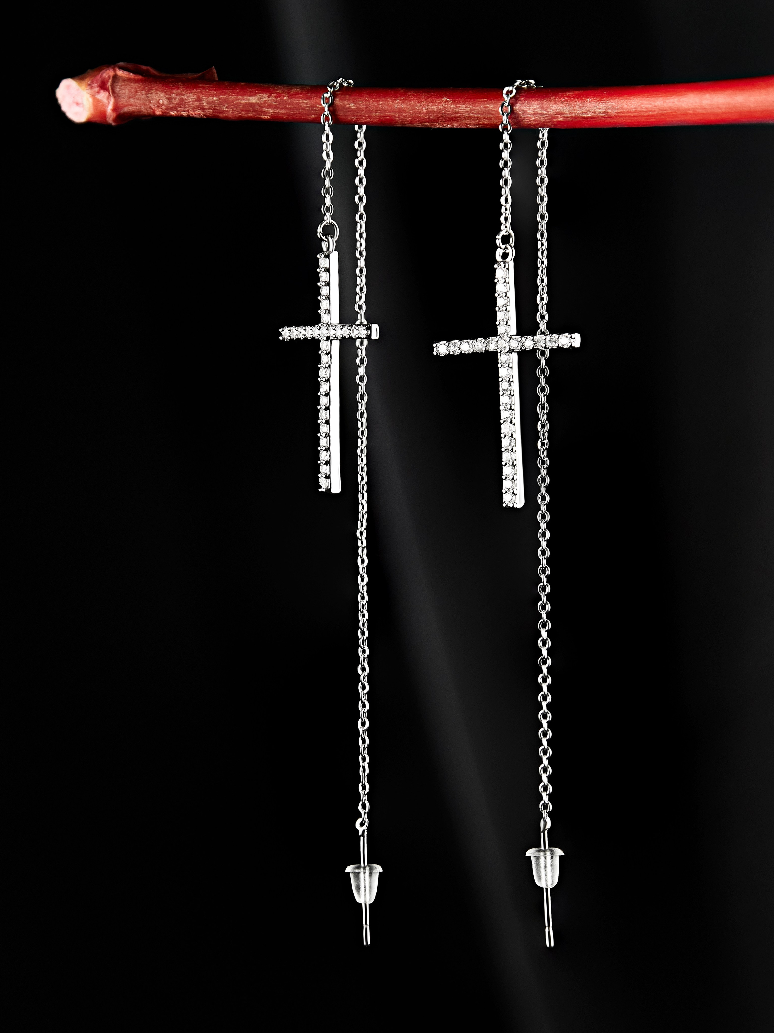 На тонкой красной веточке висят две серебряные серьги-цепочки, каждая из которых украшена крестом, инкрустированным мелкими драгоценными камнями. Серьги состоят из длинных свисающих цепочек, заканчивающихся маленькими серебряными кончиками. Фон черный, что подчеркивает детали украшения.