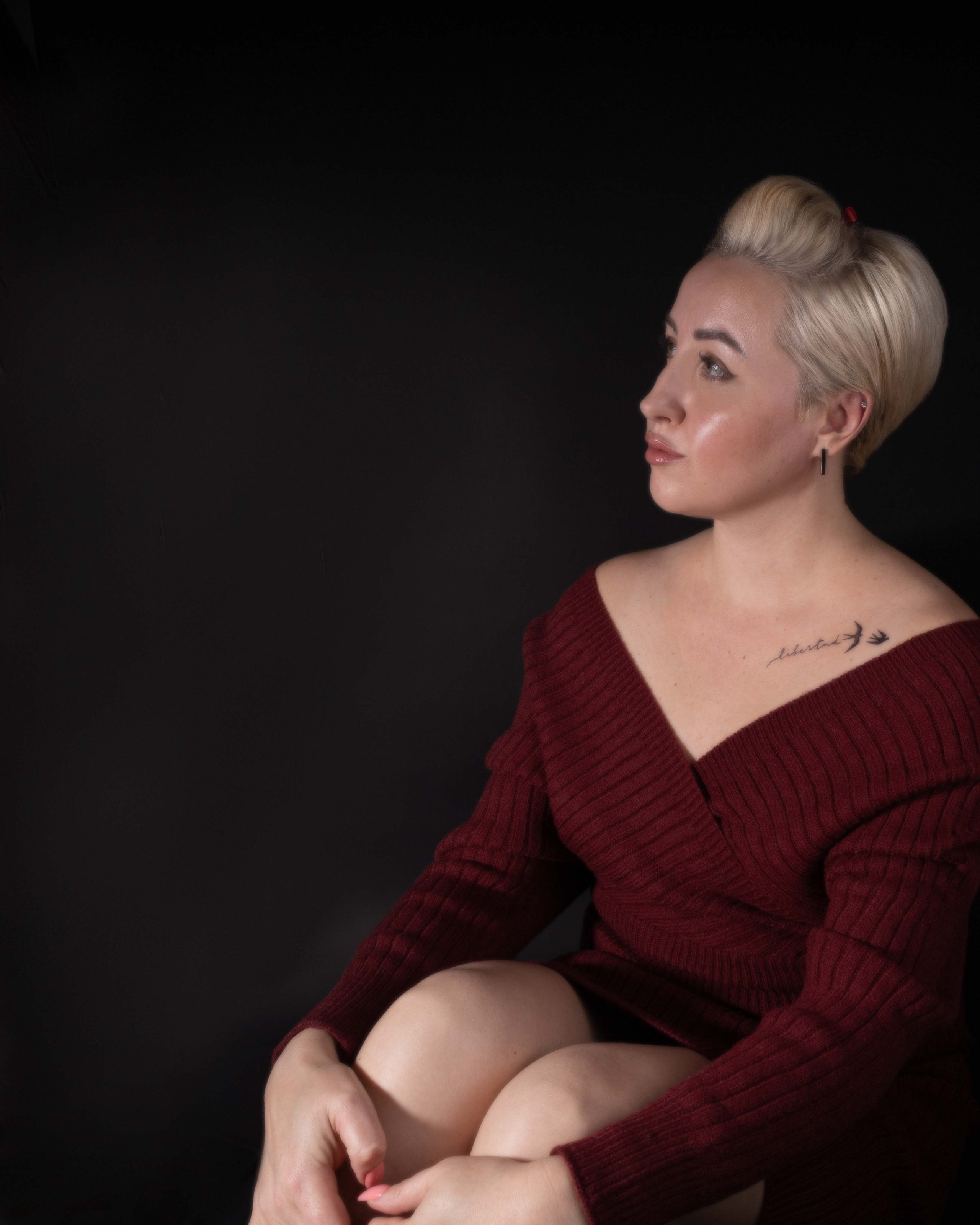 Женщина в бордовом свитере, светлые волосы, татуировка на ключице, тёмный фон