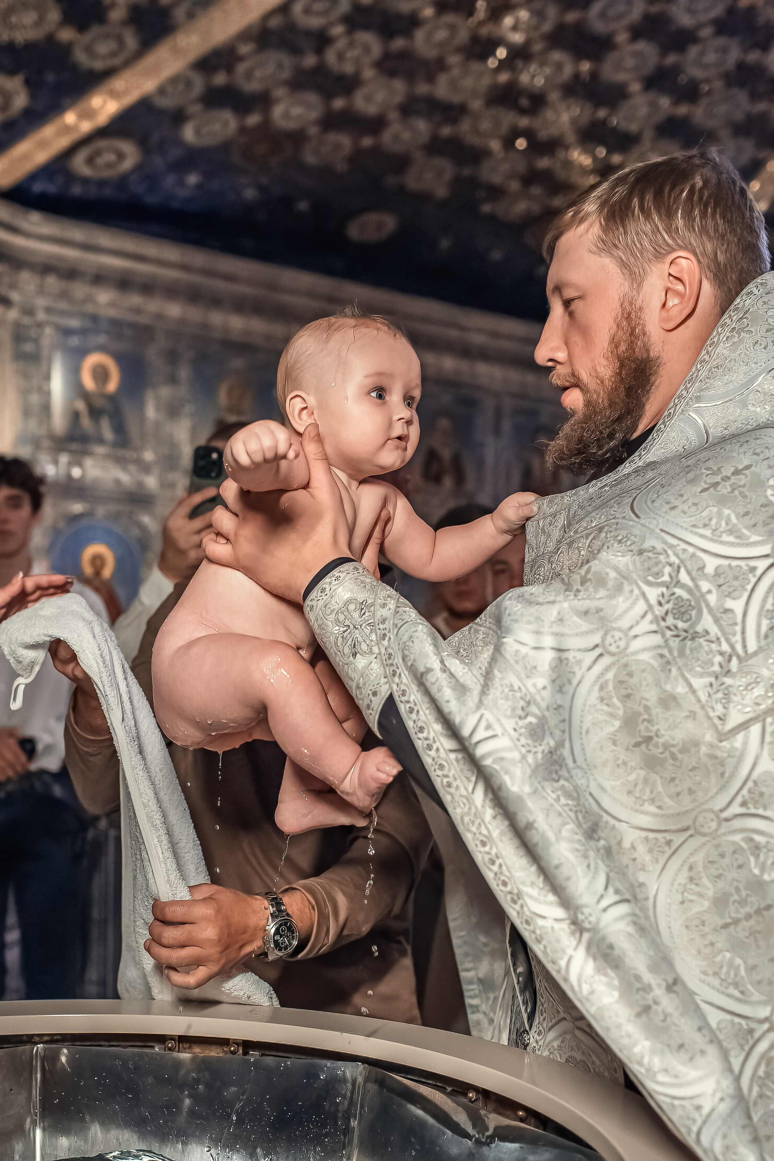 Батюшка поднимает малышку на руки из воды.