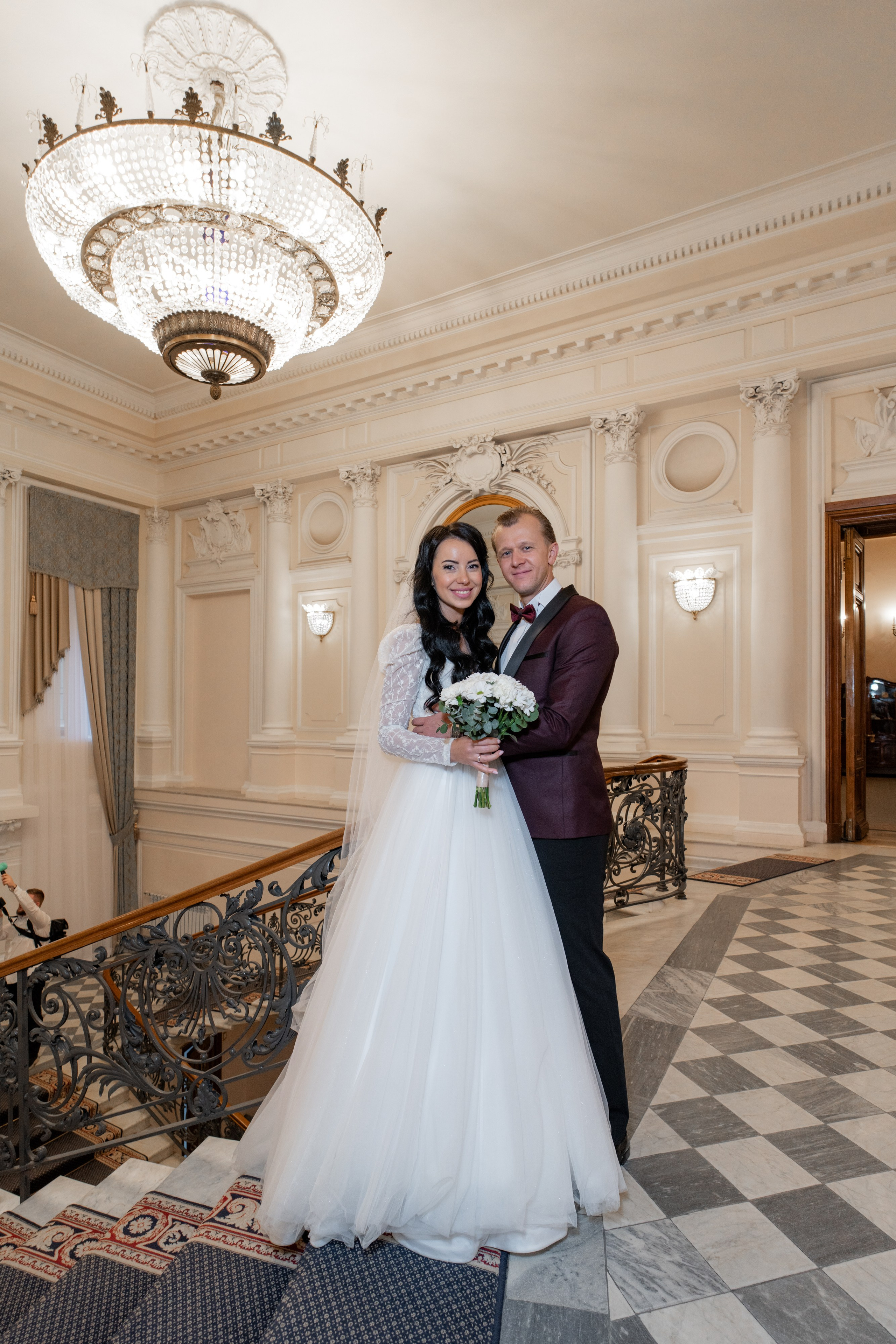 Свадебная фотосессия во дворце №2 на Фурштадской. Жених и невеста стоят на главной лестнице на фоне люстры