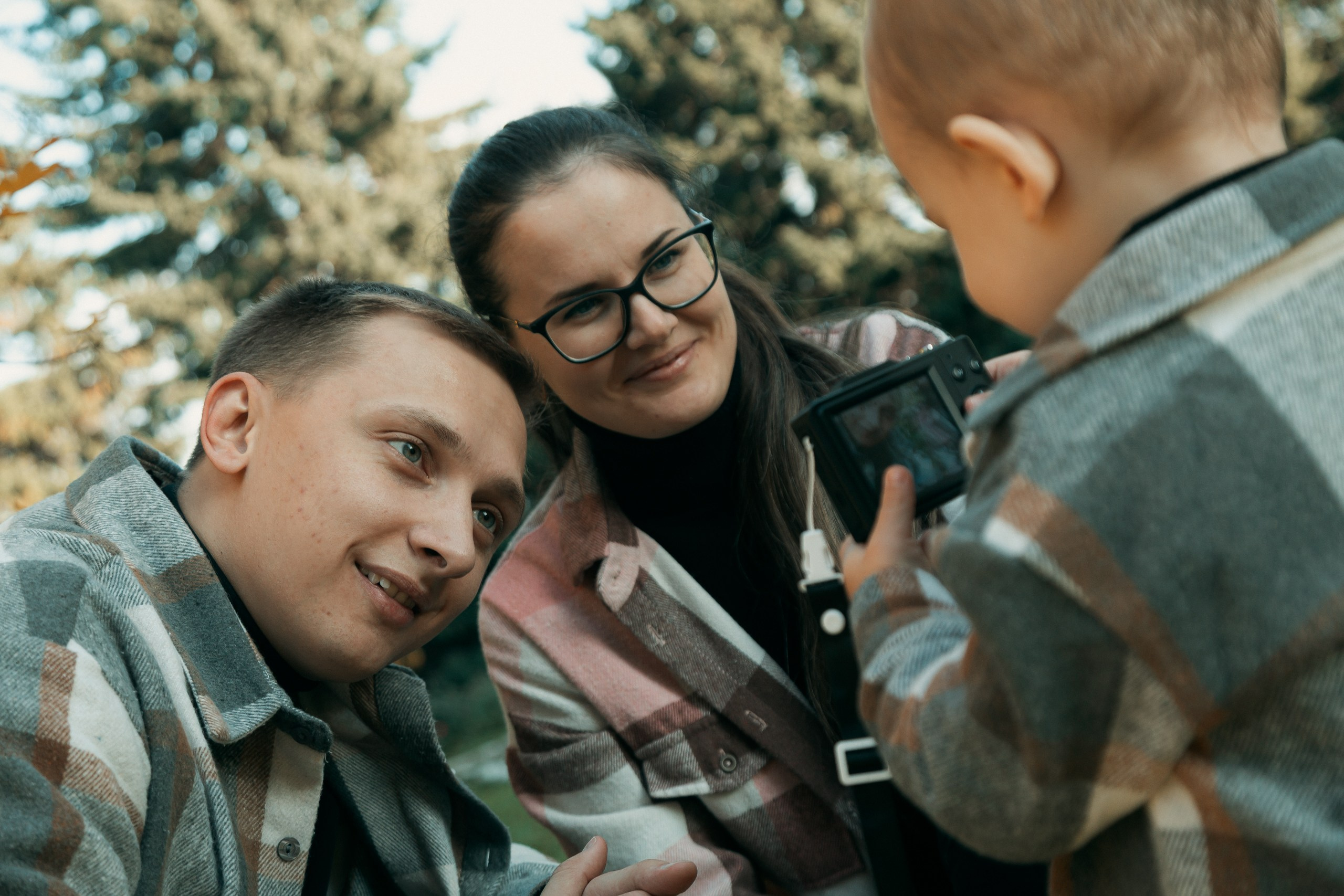 Родители и сын лежат на траве, ребёнок держит камеру, семья смеётся и рассматривает снимки.