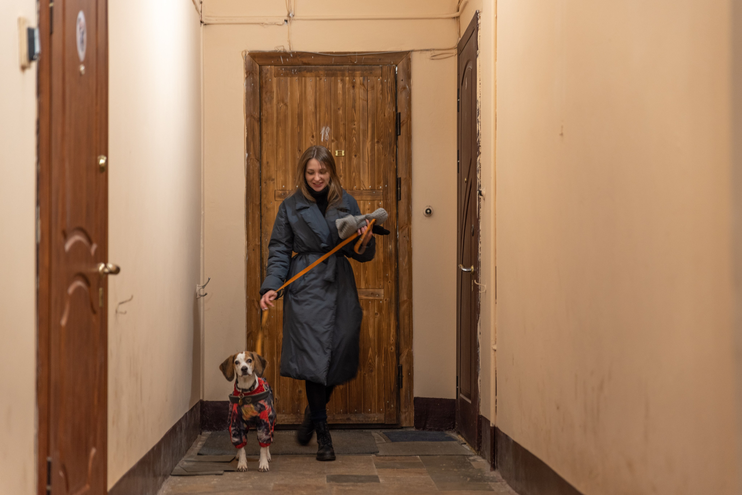 Девушка с собакой идёт по коридору. На девушке длинная куртка, за спиной дверь в квартиру.