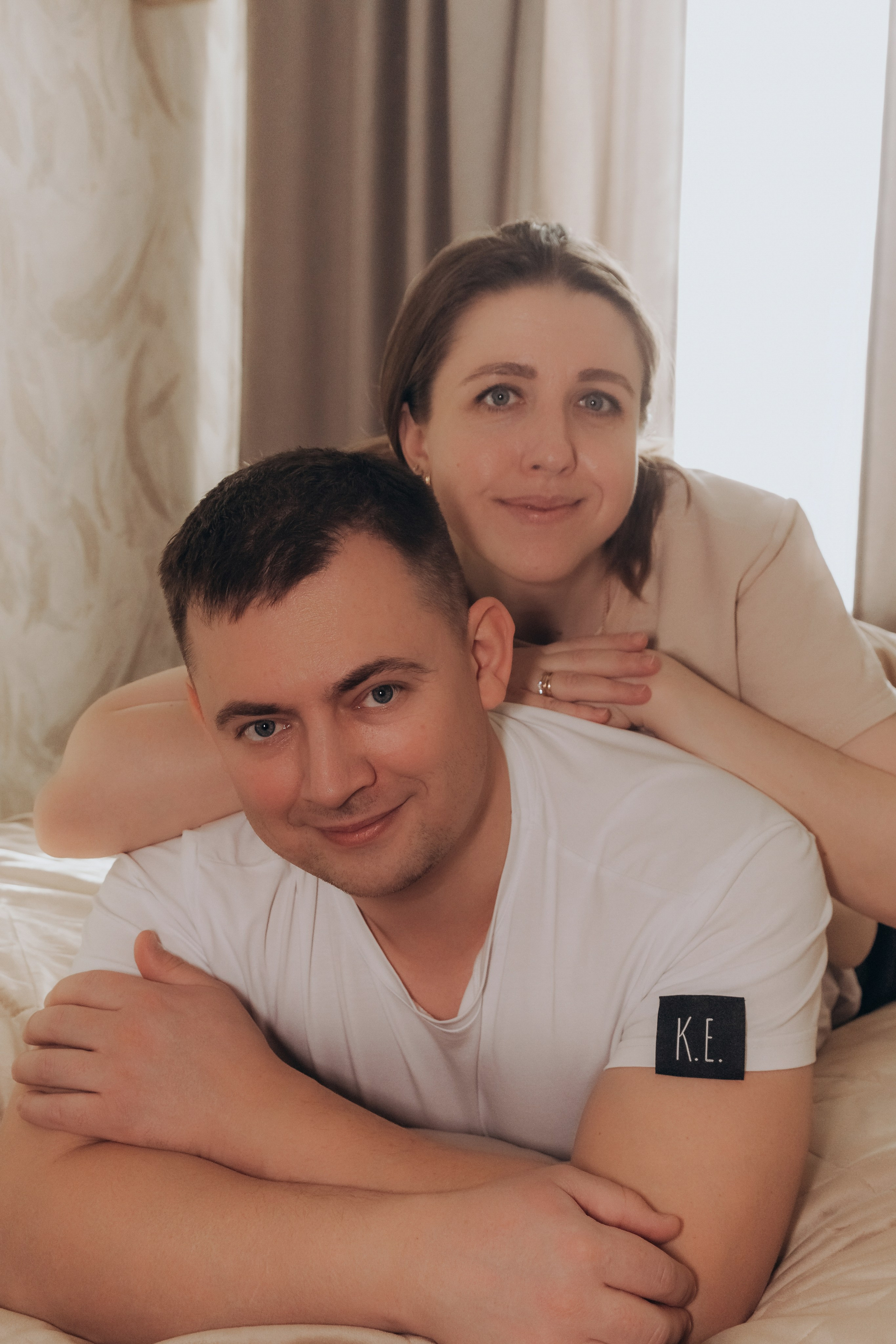Папа лежит на кровати, мама обнимает его сзади, оба улыбаются, семейный портрет.