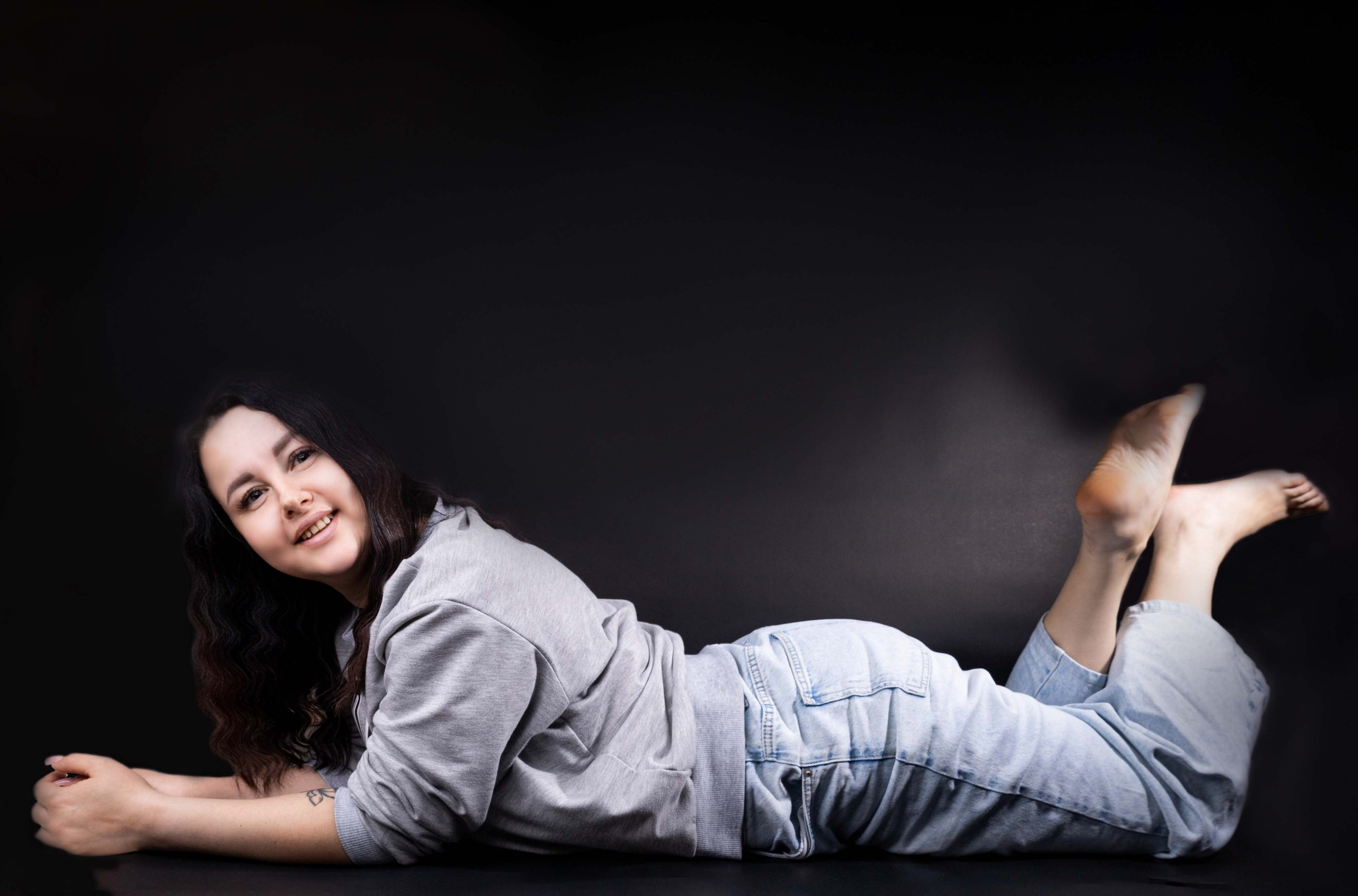 Портретная фотография молодой женщины в расслабленной позе: она лежит на животе, опираясь на руки, в повседневной одежде — свитшоте и джинсах. Фон однотонный, тёмный