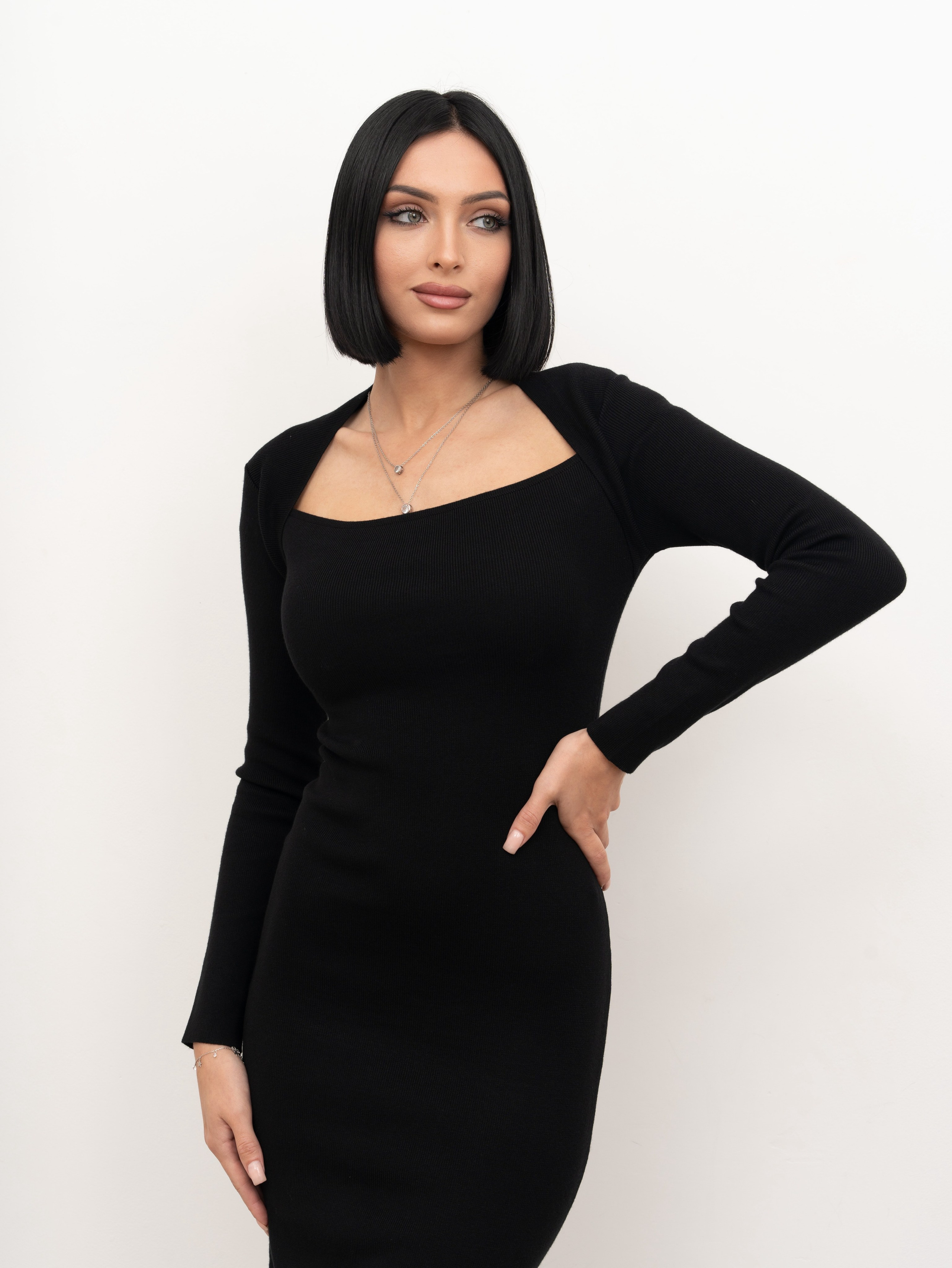 Модель в черном платье футляр и украшением на шее стоит на белом фоне