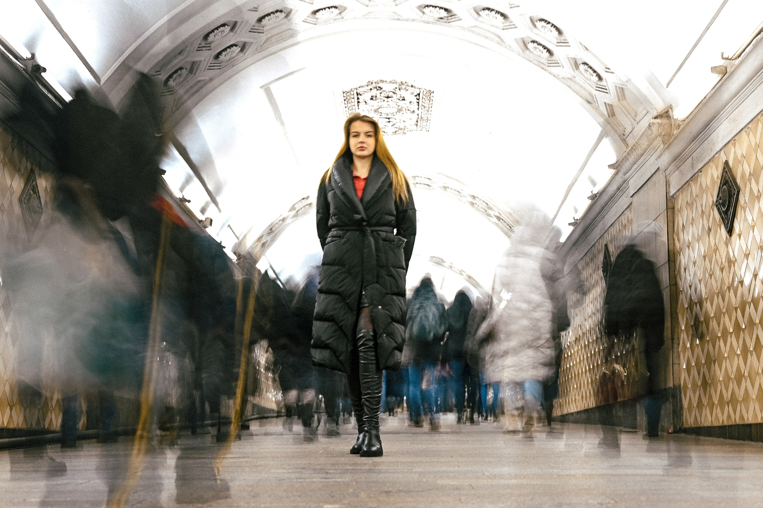 Молодая женщина в темном пуховике и высоких сапогах стоит на одной из станций метро. Она имеет длинные волосы, которые свободно спадают по плечам. Вокруг нее движутся люди, создавая эффект размытия на переднем плане, что акцентирует внимание на модели. Станция оформлена в классическом стиле с арочными сводами и светильниками, придавая ей элегантность и исторический характер. Освещение яркое, подчеркивающее детали интерьера и образ женщины. Фотография передает динамику городской жизни и контраст между спокойствием модели и движением вокруг нее.