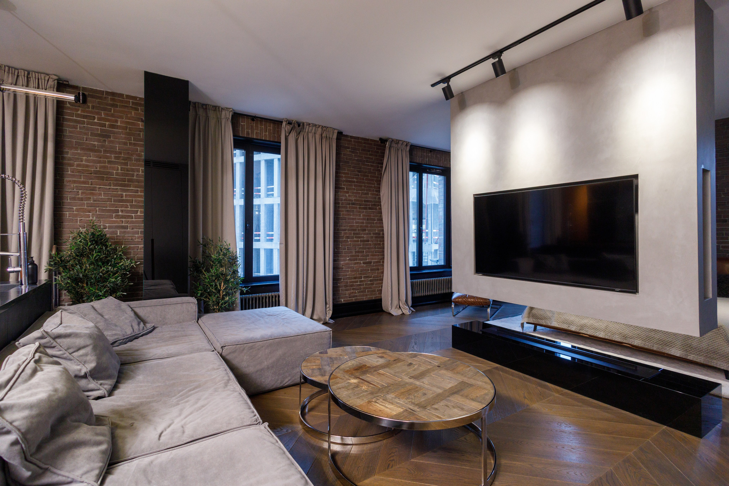 Современная гостиная в стиле лофт с кирпичными стенами, большим угловым диваном, деревянным паркетом «ёлочкой», круглыми журнальными столиками и телевизором на минималистичной перегородке с трековым освещением.