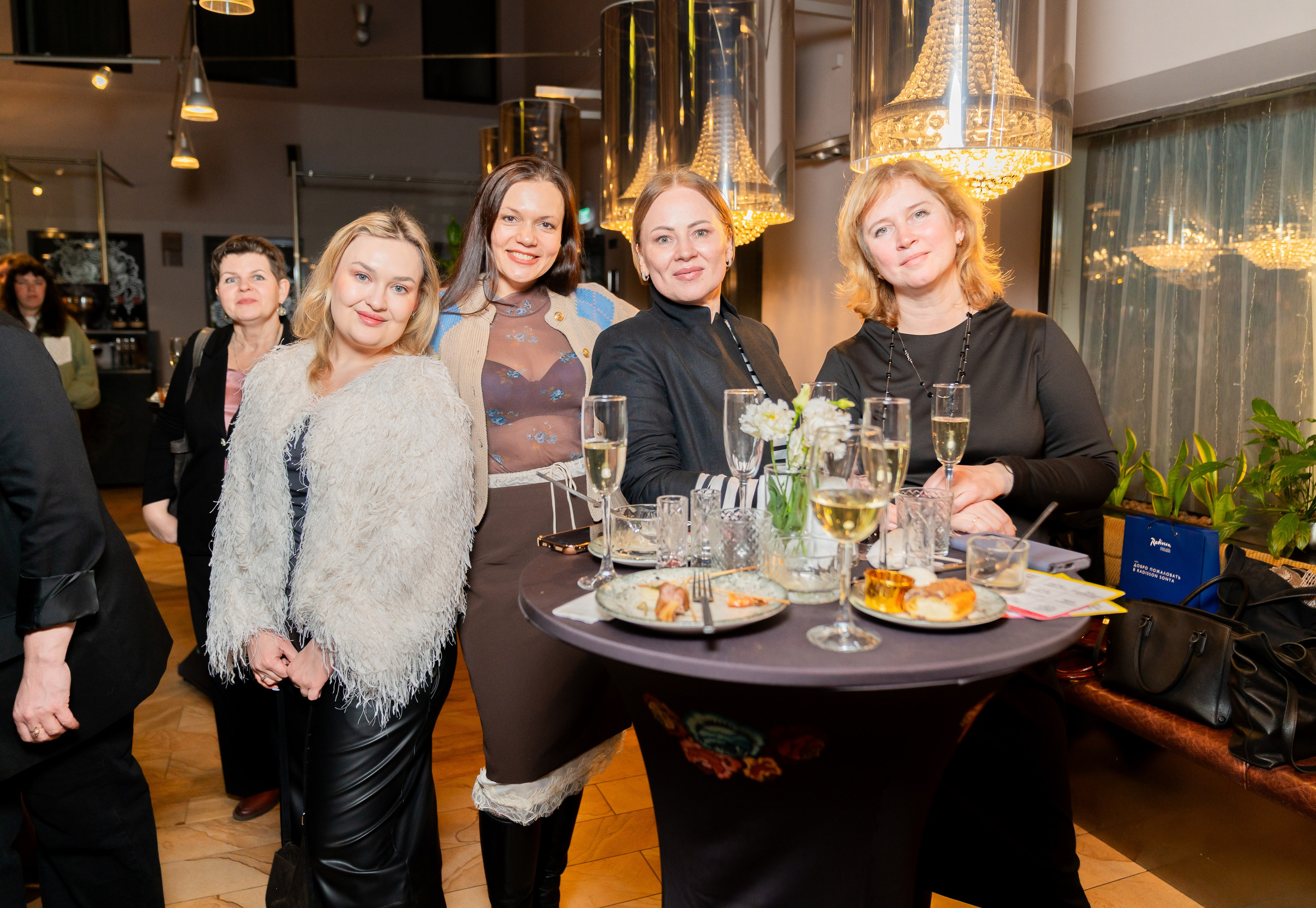 Четыре красивые женщины позируют за праздничным столом с шампанским и закусками на мероприятии в Radisson Sonya. Фотография Евгения Мальцева (evrednix.ru).