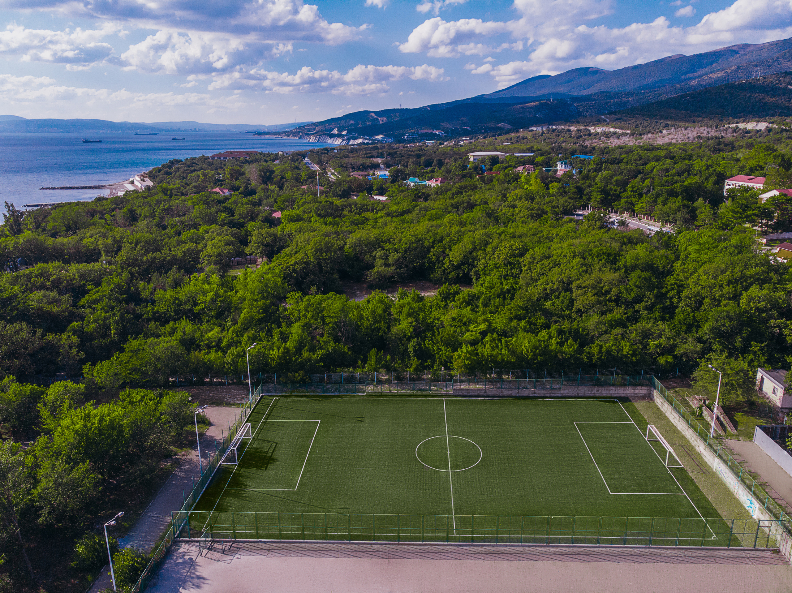 Вид на футбольное поле с высоты, вокруг поля зеленый лес, вдали видно черное море и горы.