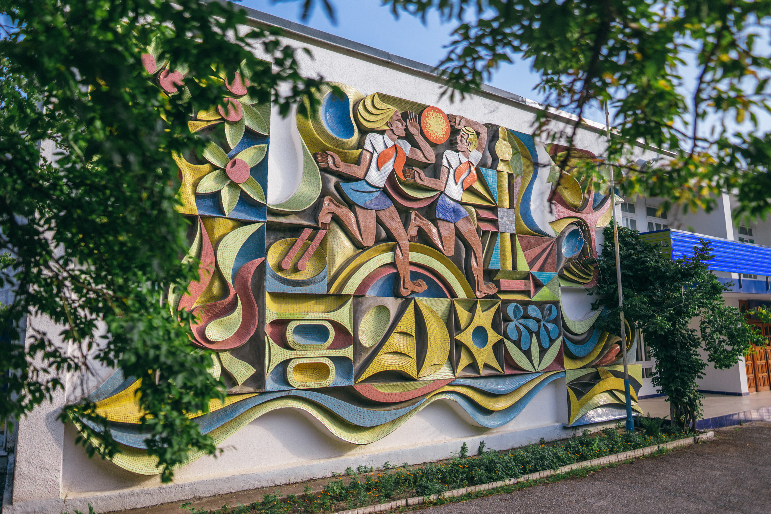 Мозаика на стене кают-компании, ветви деревьев с зелеными листьями на переднем плане, мальчик и девочка на панно тянутся к солнцу.