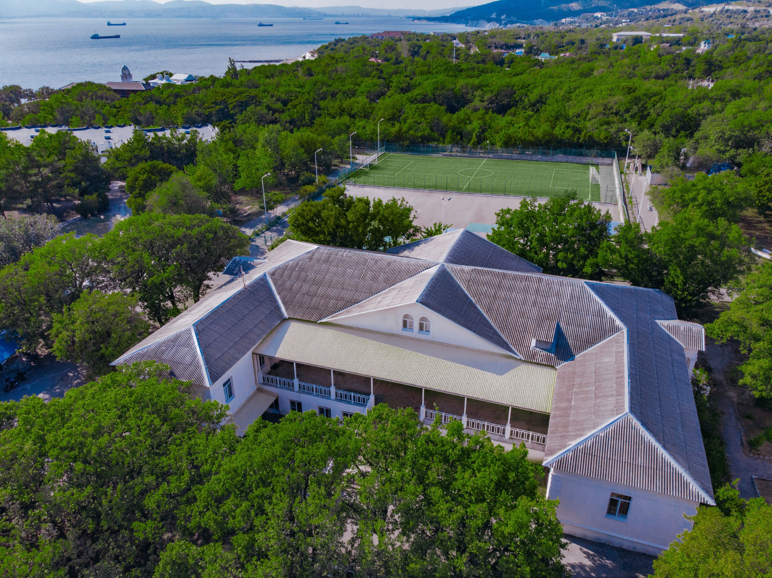 Вид с воздуха на детский лагерь, на первом плане здание корпуса Ледокол, на втором плане спортивная площадка с зеленым покрытием. На заднем плане горы и Черное море.
