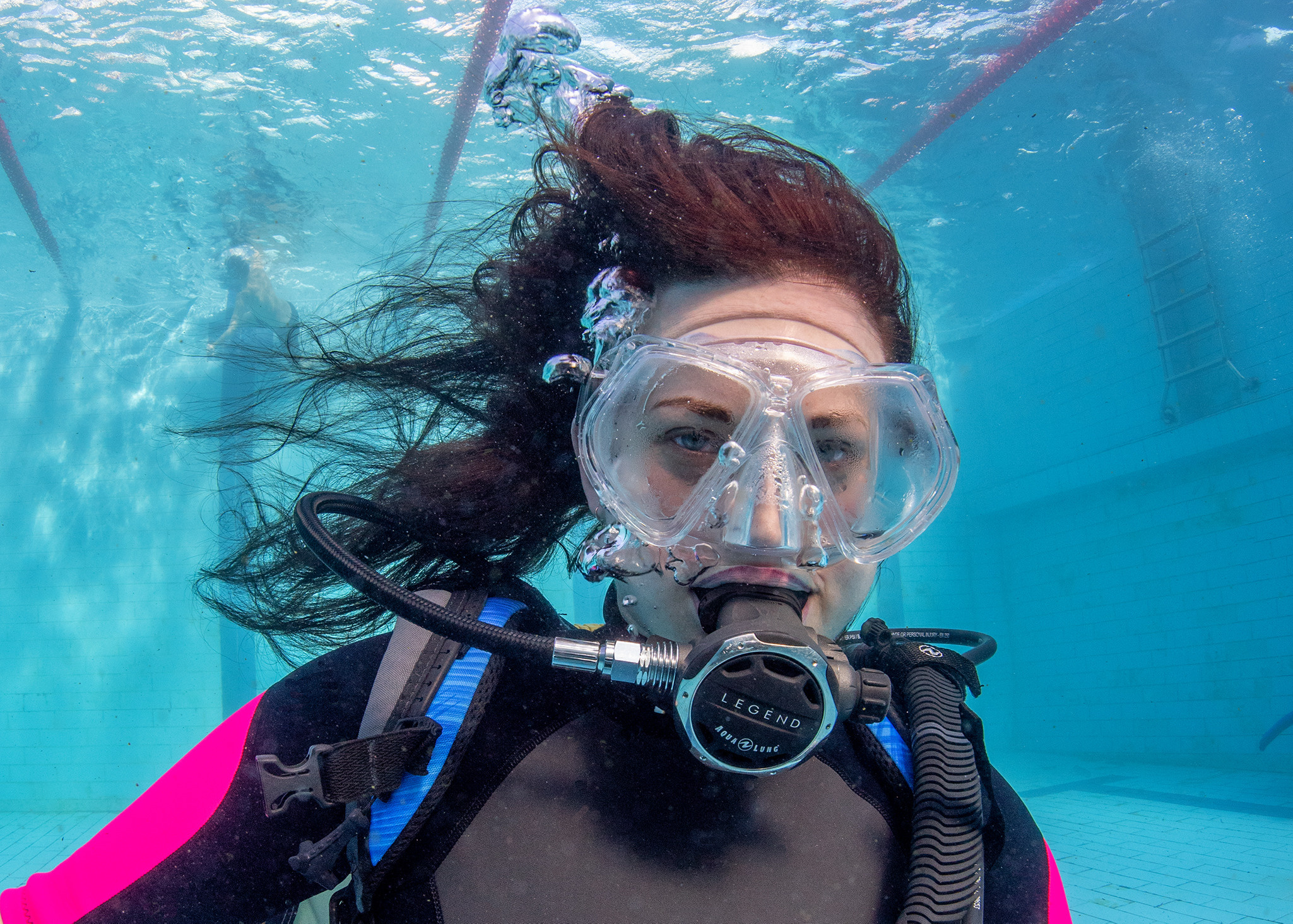 Красивая девушка дайвер под водой в бассейне смотрит в камеру фотографа