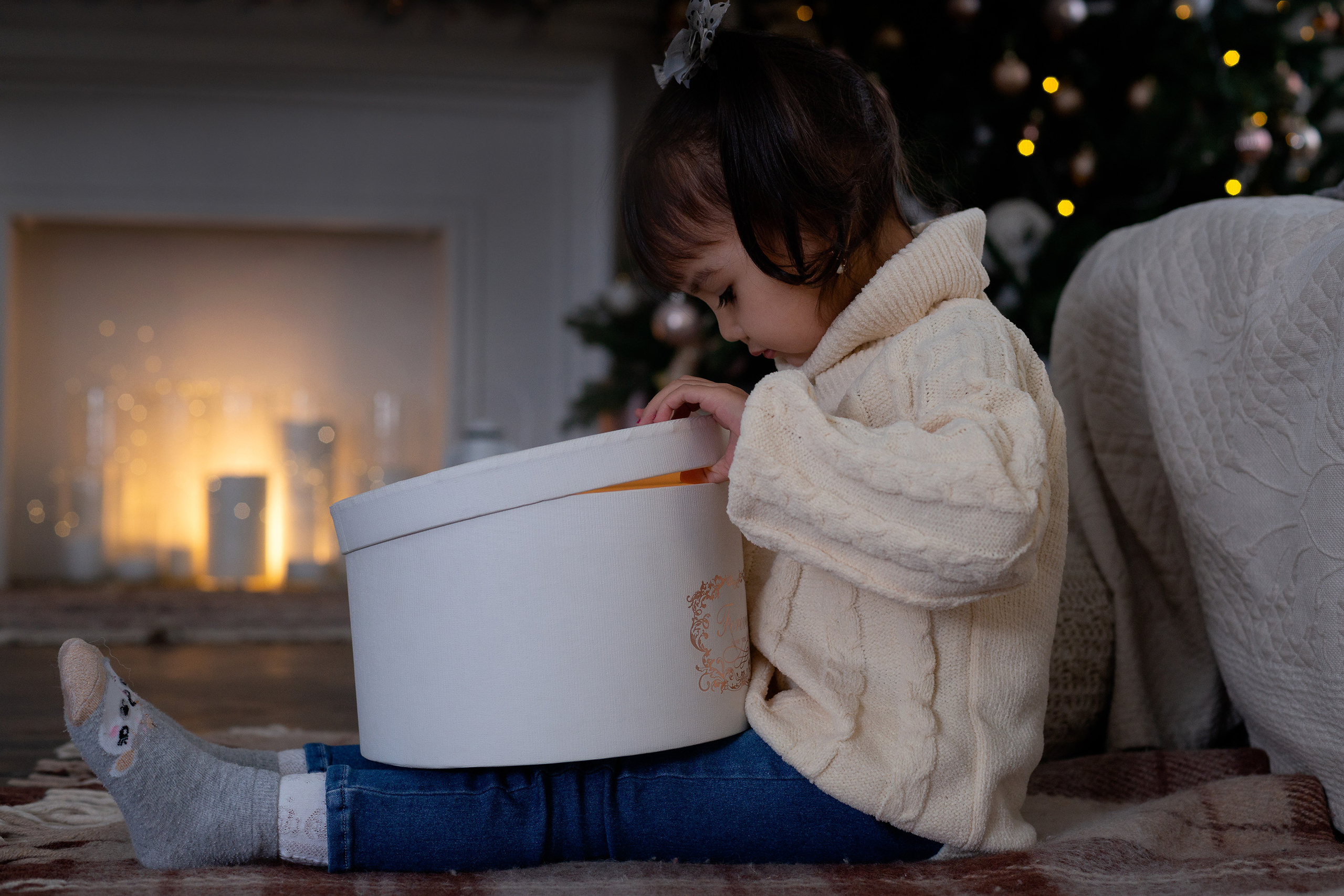 Маленькая девочка сидя на полу открывает новогодний подарок, а на заднем фоне горящий камин и елка с гирляндами.