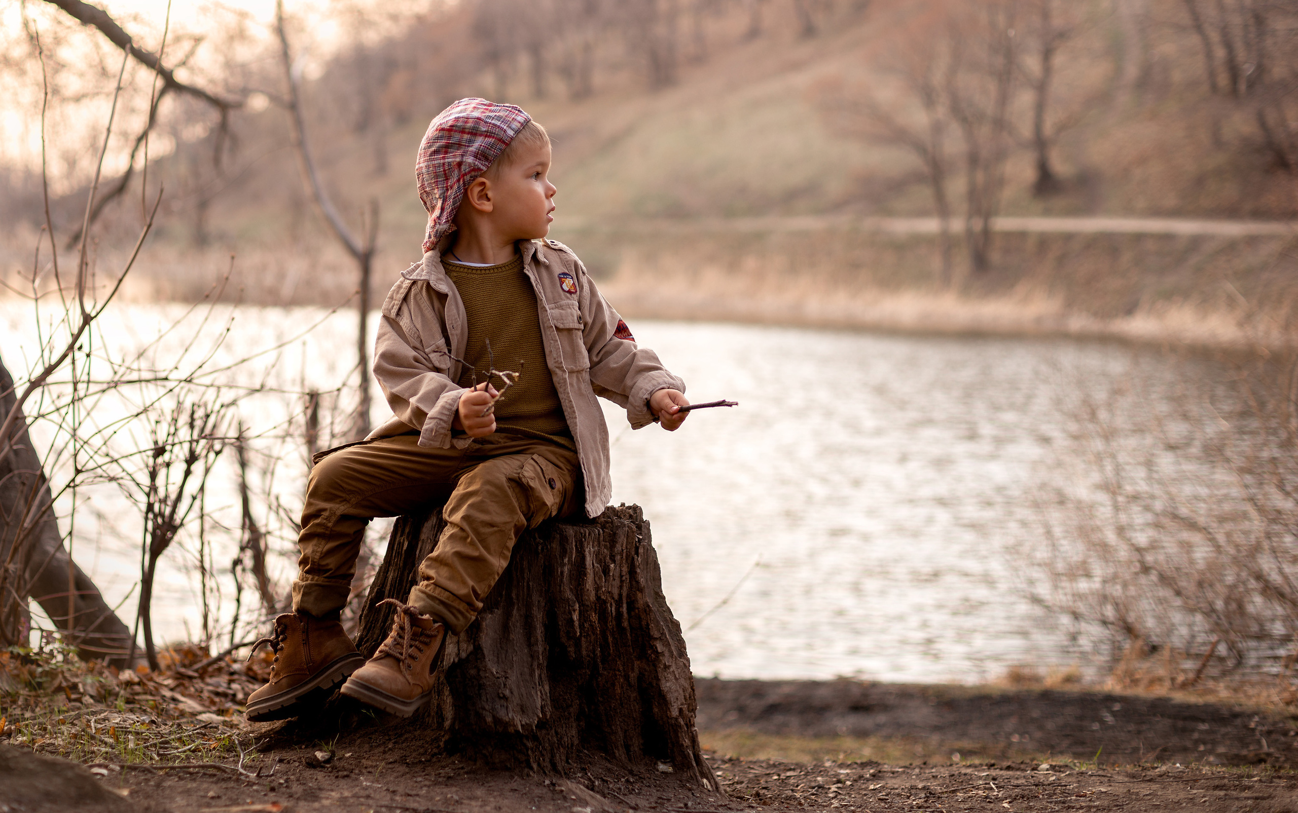 Маленький мальчик сидит на пеньке около озера и смотрит вдаль.