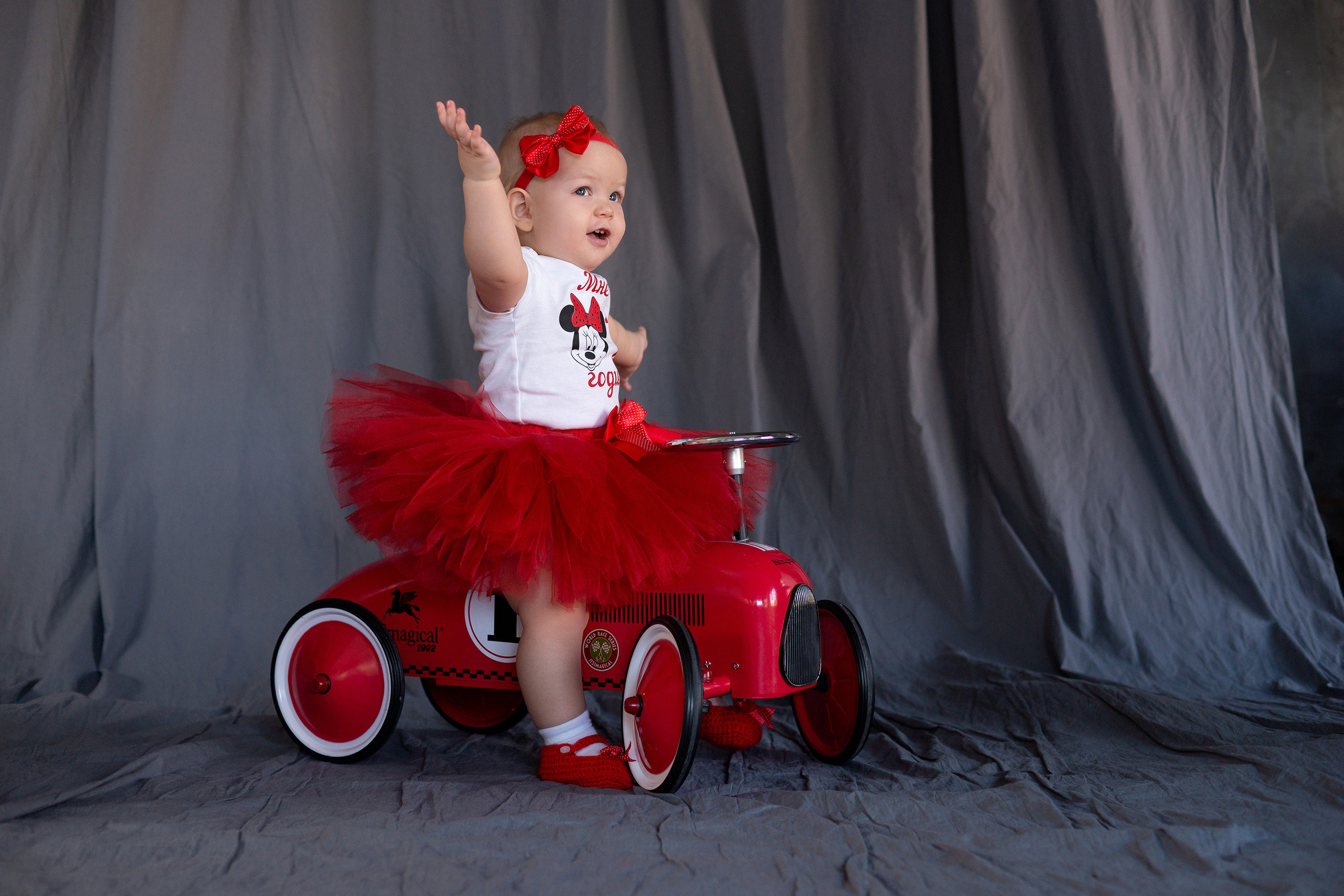 Маленькая девочка в красной юбке сидит на красной машинке и машет кому-то рукой смотря в сторону.