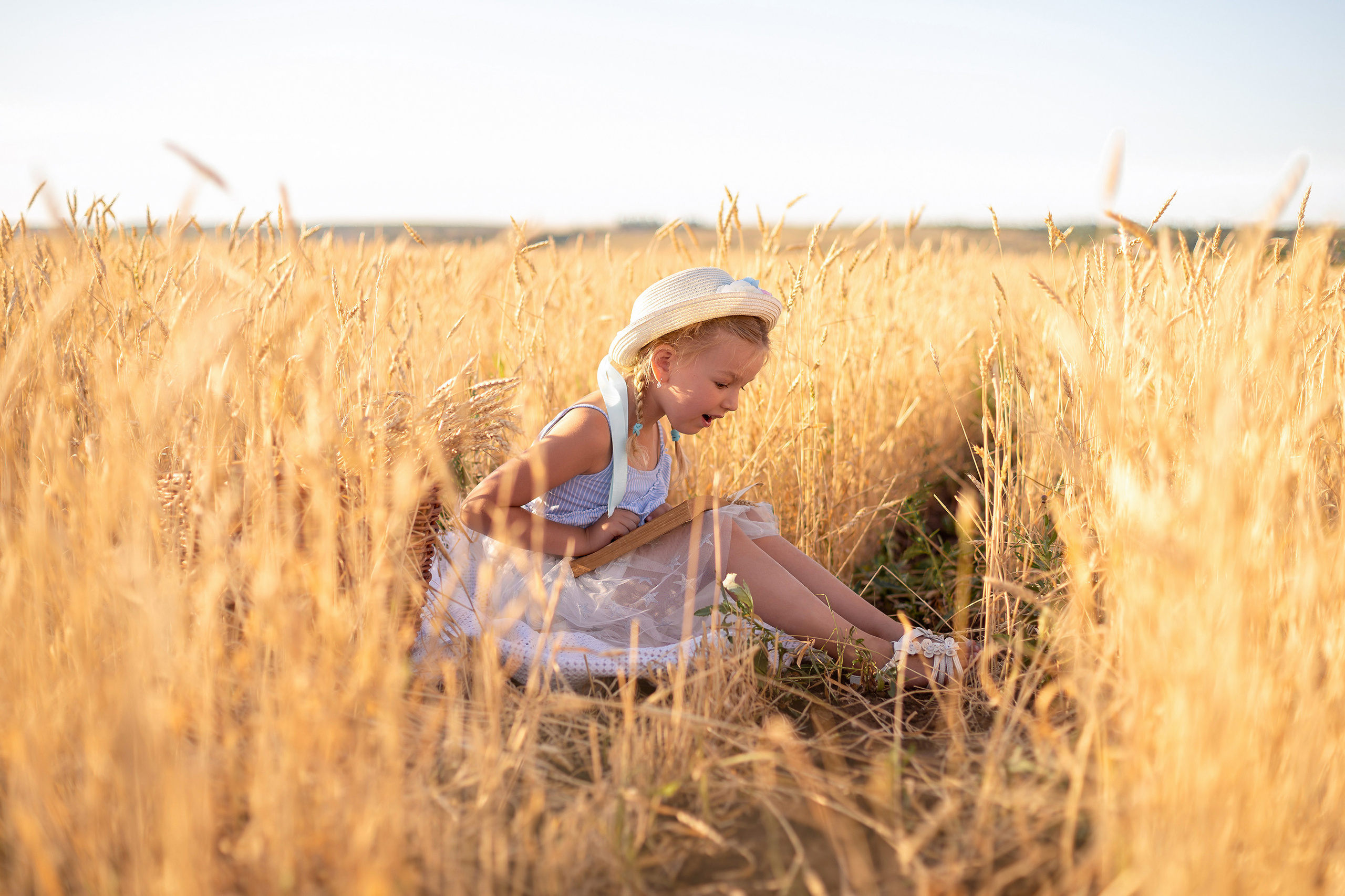 Маленькая девочка с шляпкой на голове, и книжкой на коленках, смотрит на свои ноги сидя посередине пшеничного поля.