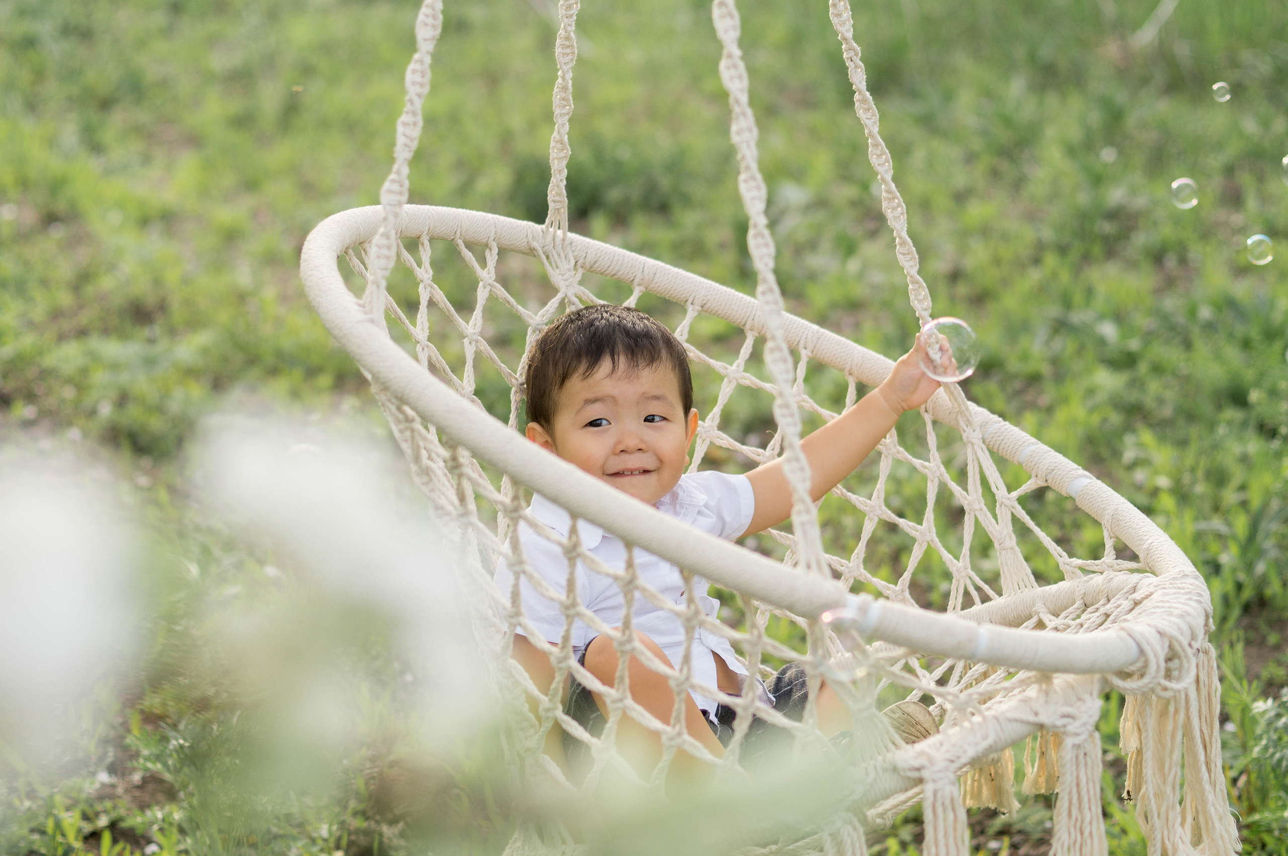 Маленький темный мальчик сидит на подвесном кресле и смотрит в камеру на фоне зеленой лужайки.