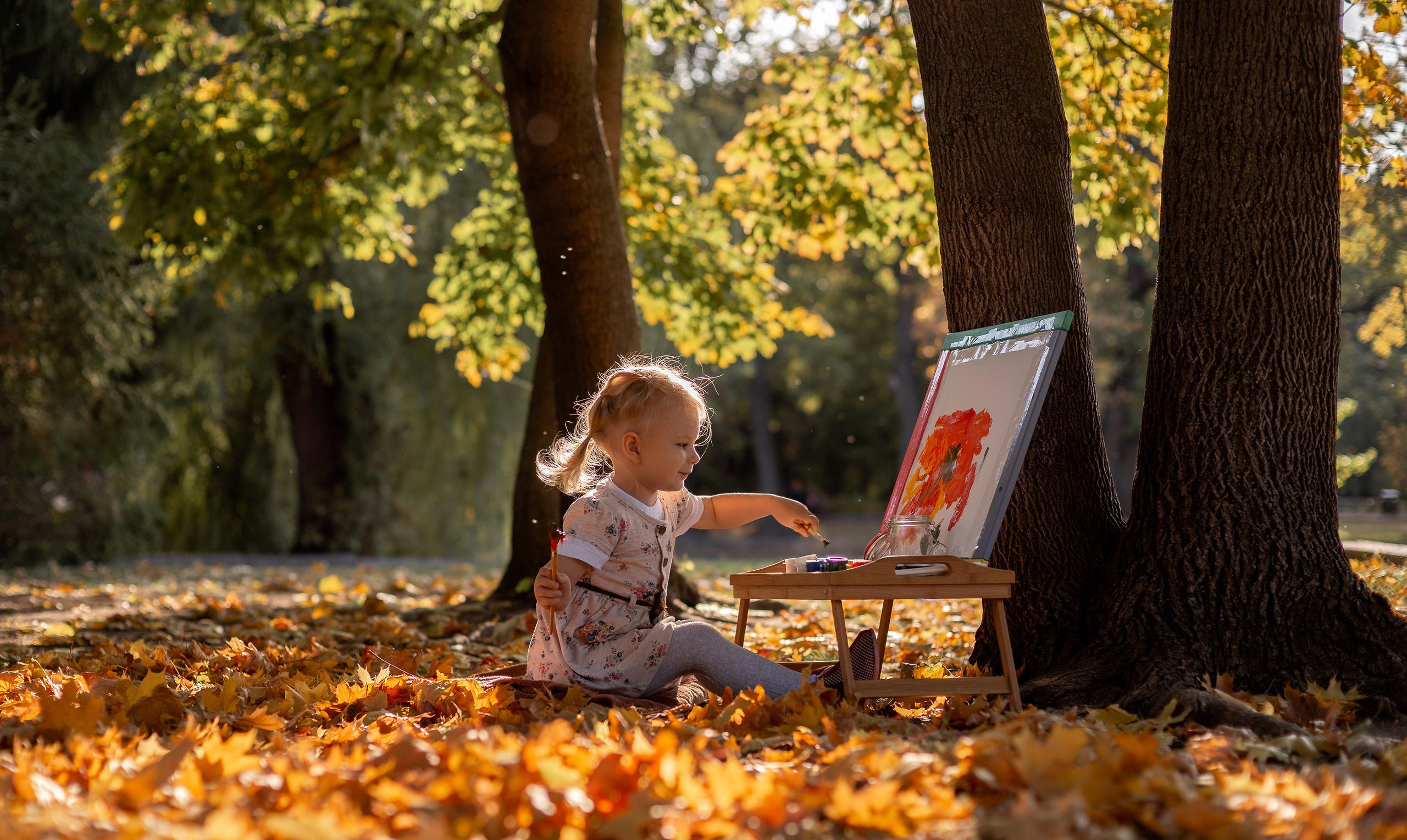 Меленькая девочка сидит в осеннем парке и рисует на мольберте, рядом три дуба и желтые листья вокруг.
