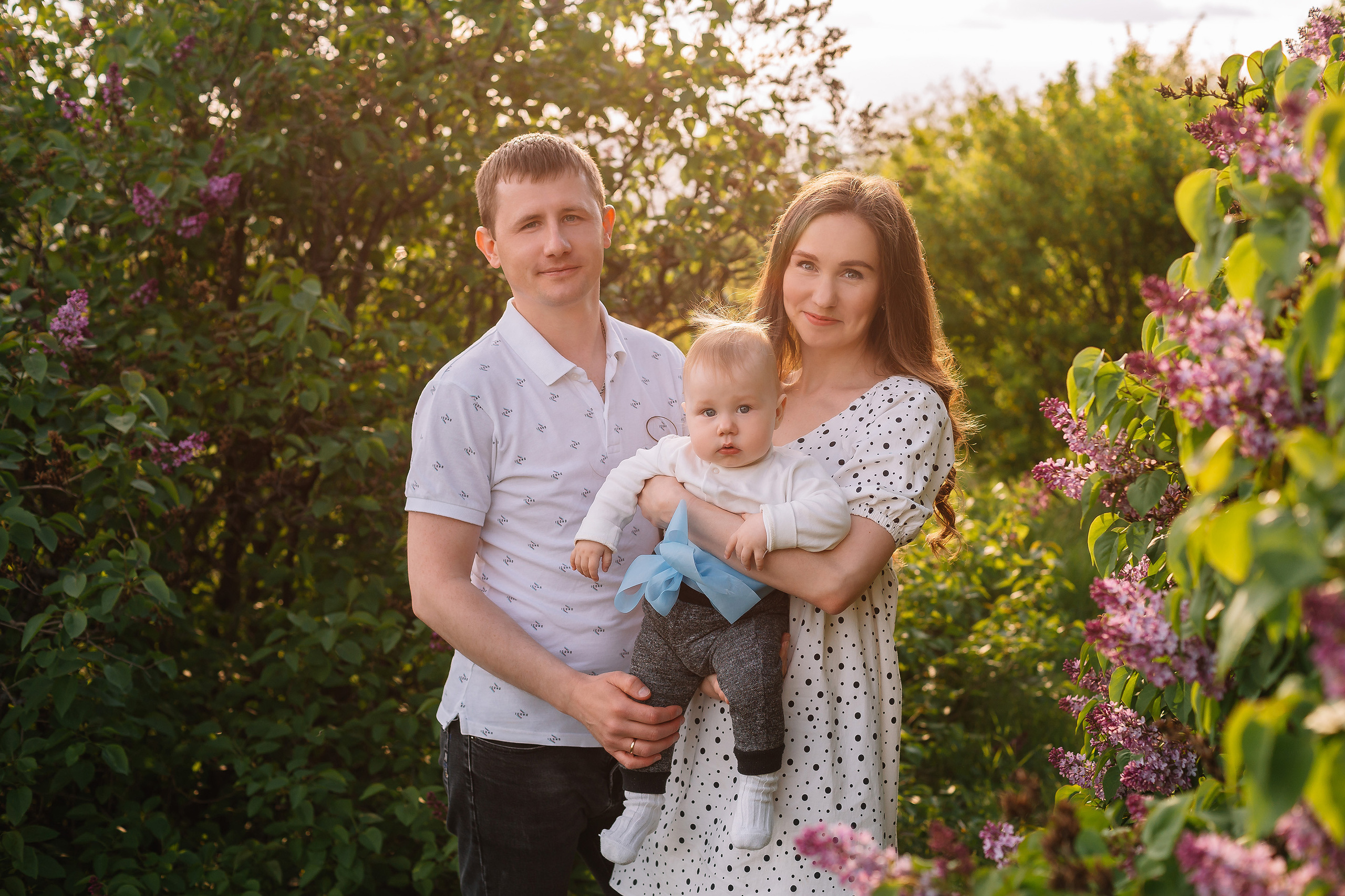 Красивая пара с маленьким ребенком стоит подсвеченная сзади лучами солнца около куста сирени.