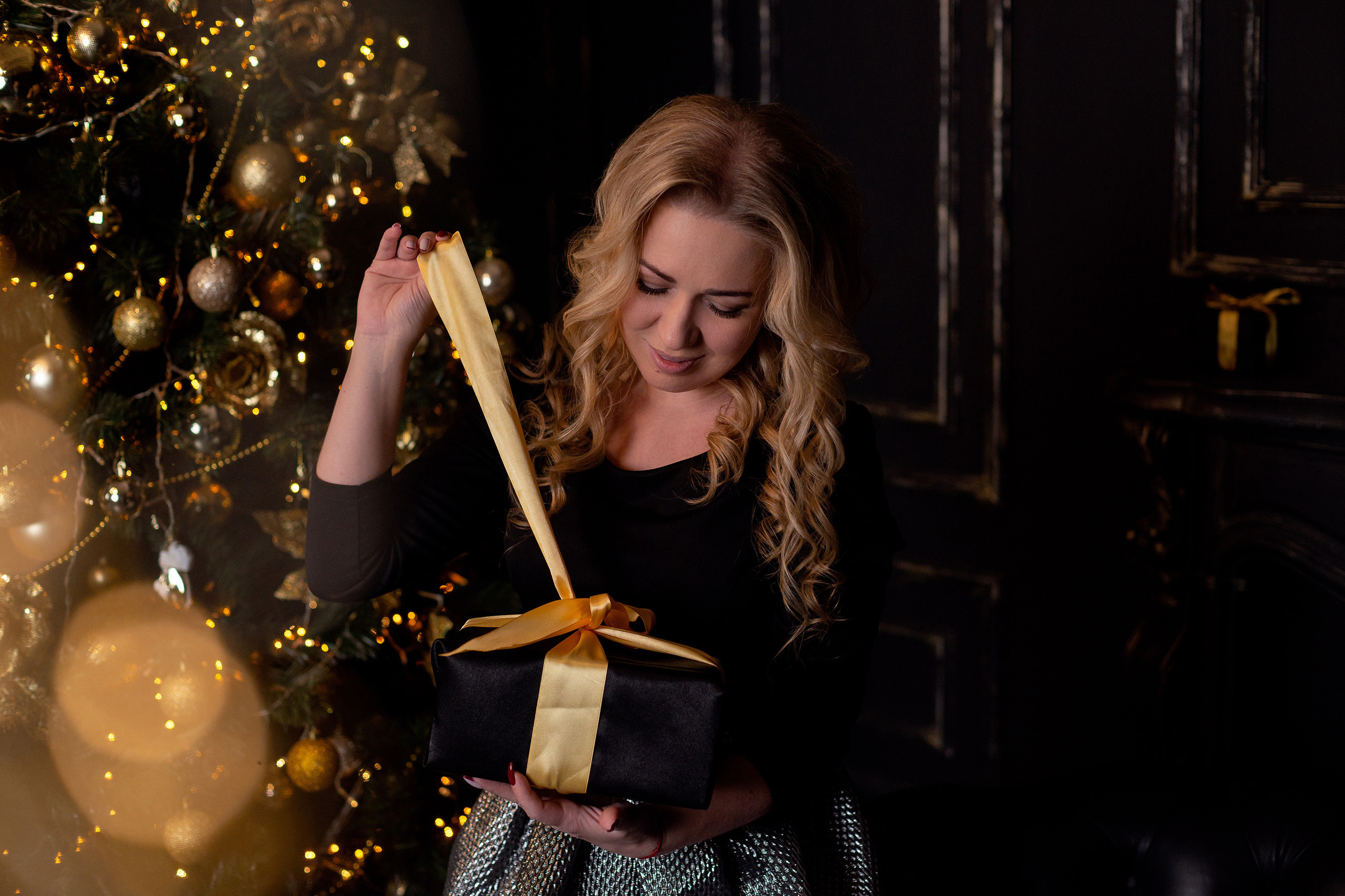 Девушка в черной блузке распаковывает новогодний подарок на фоне гирлянд.