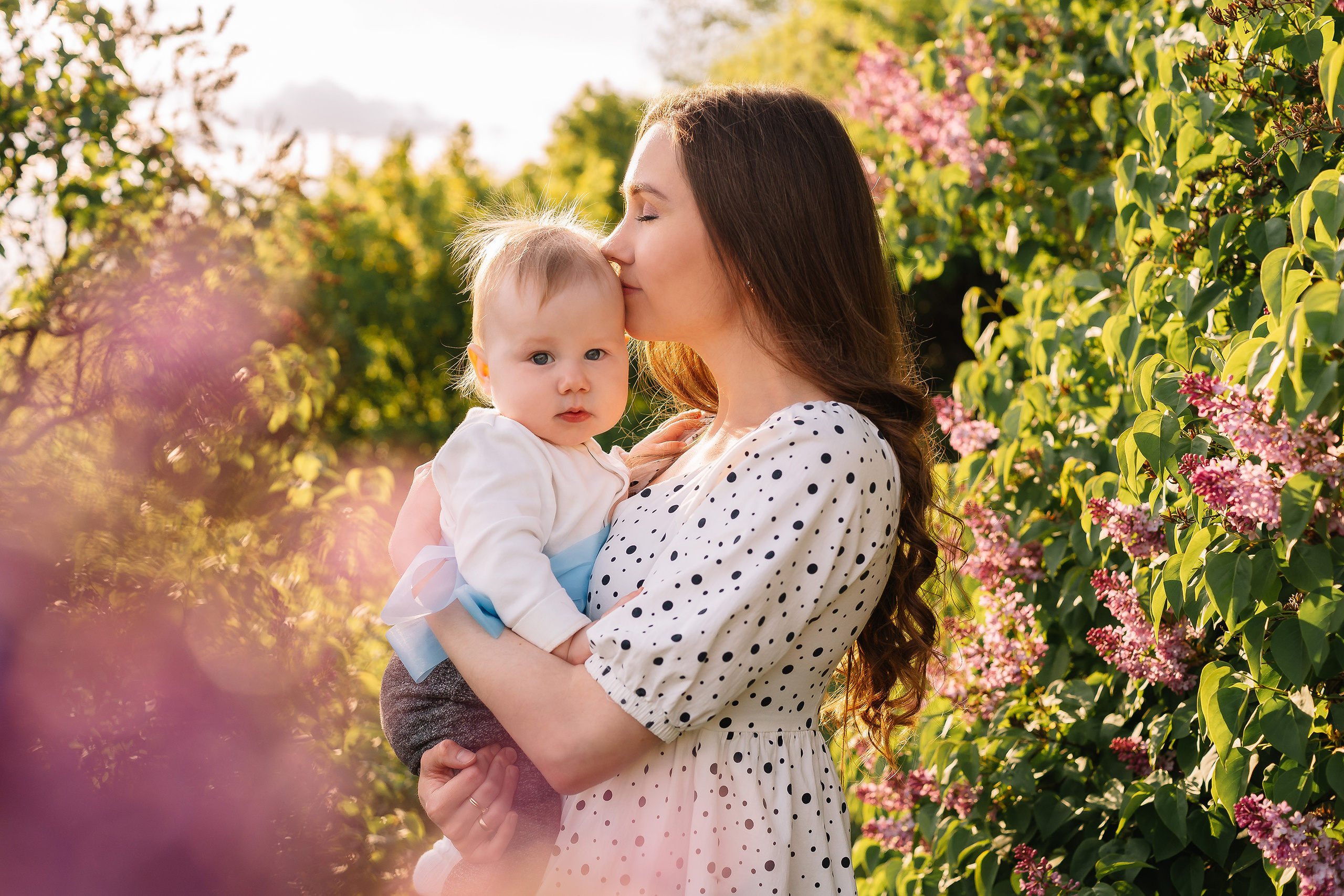 Мать в белом платье в горошек целует маленького сына на руках, а сын мило смотрит в камеру. Рядом кусты сирени.
