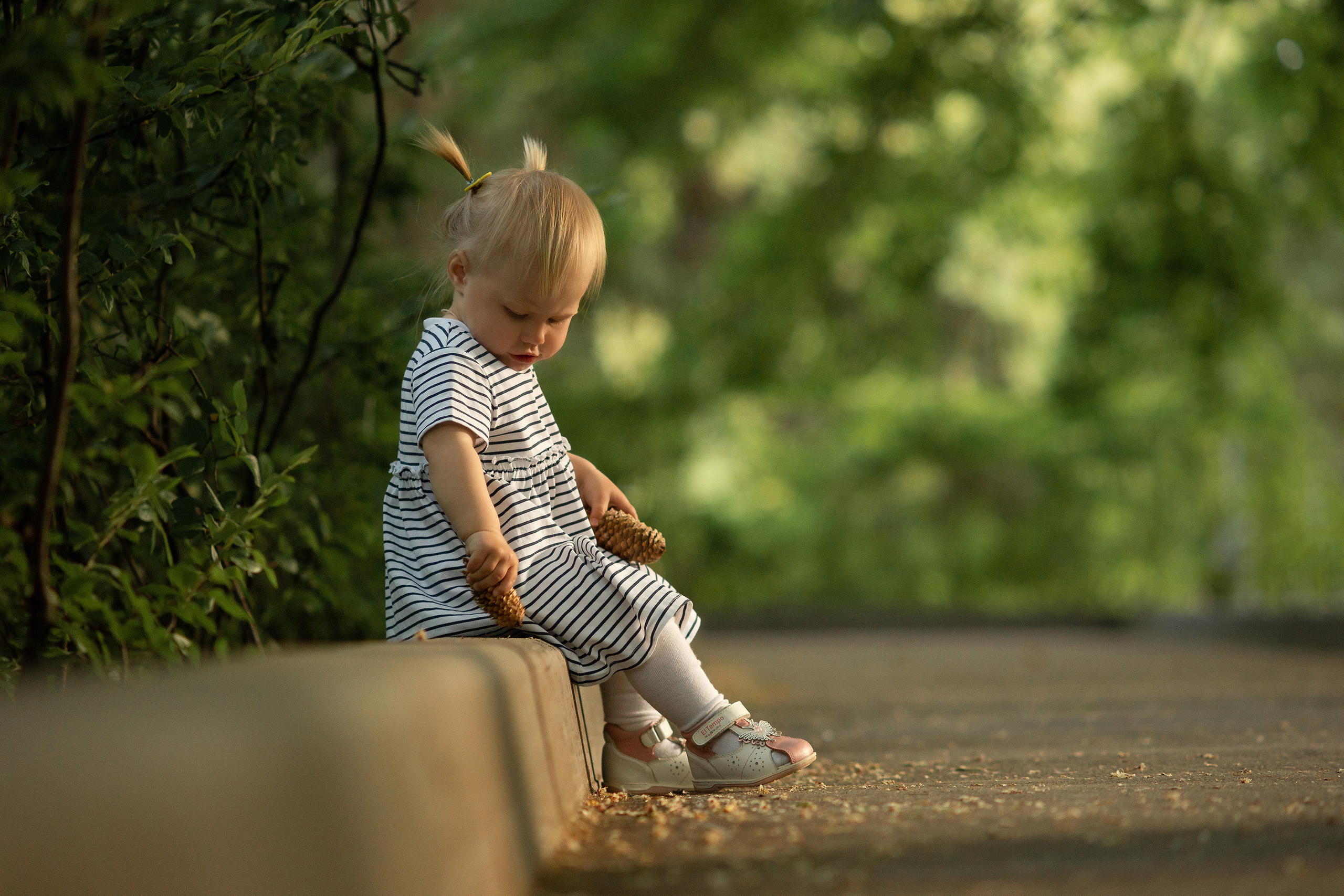 Маленькая девочка в легком платье сидит на бордюре и играет с шишкой на аллее горпарка Саратова.