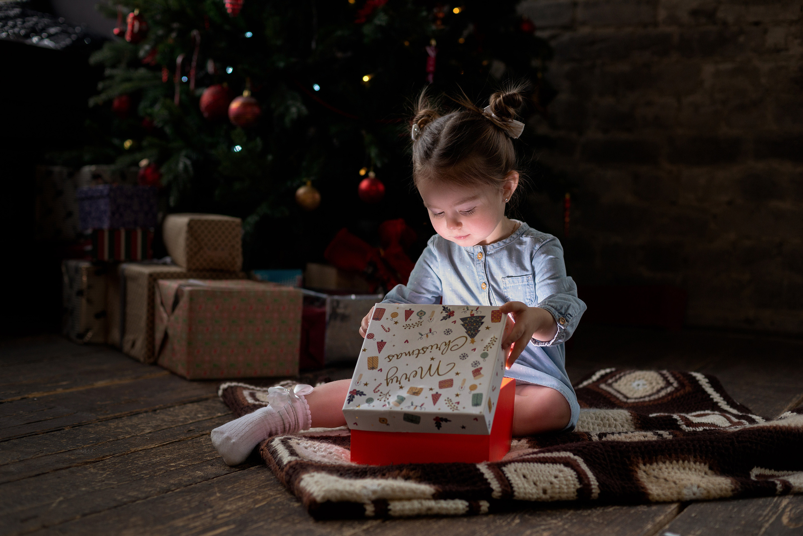 Маленькая девочка с бантиками на голове заглядывает в подарочную новогоднюю коробку, а из коробки по комнате разливается свет освещая её лицо.