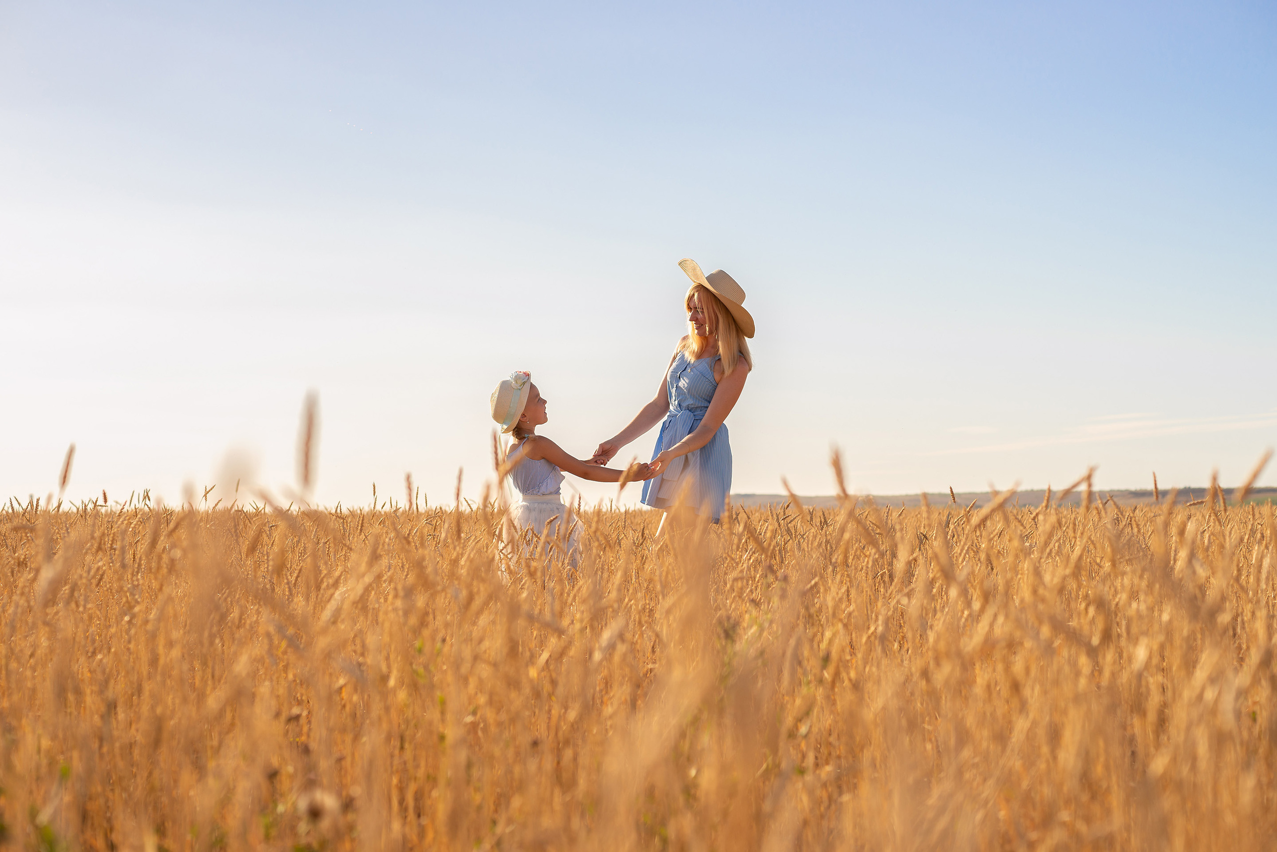 Мама и дочка взявшись за руки смотрят друг на друга в пшеничном поле на фоне голубого неба.