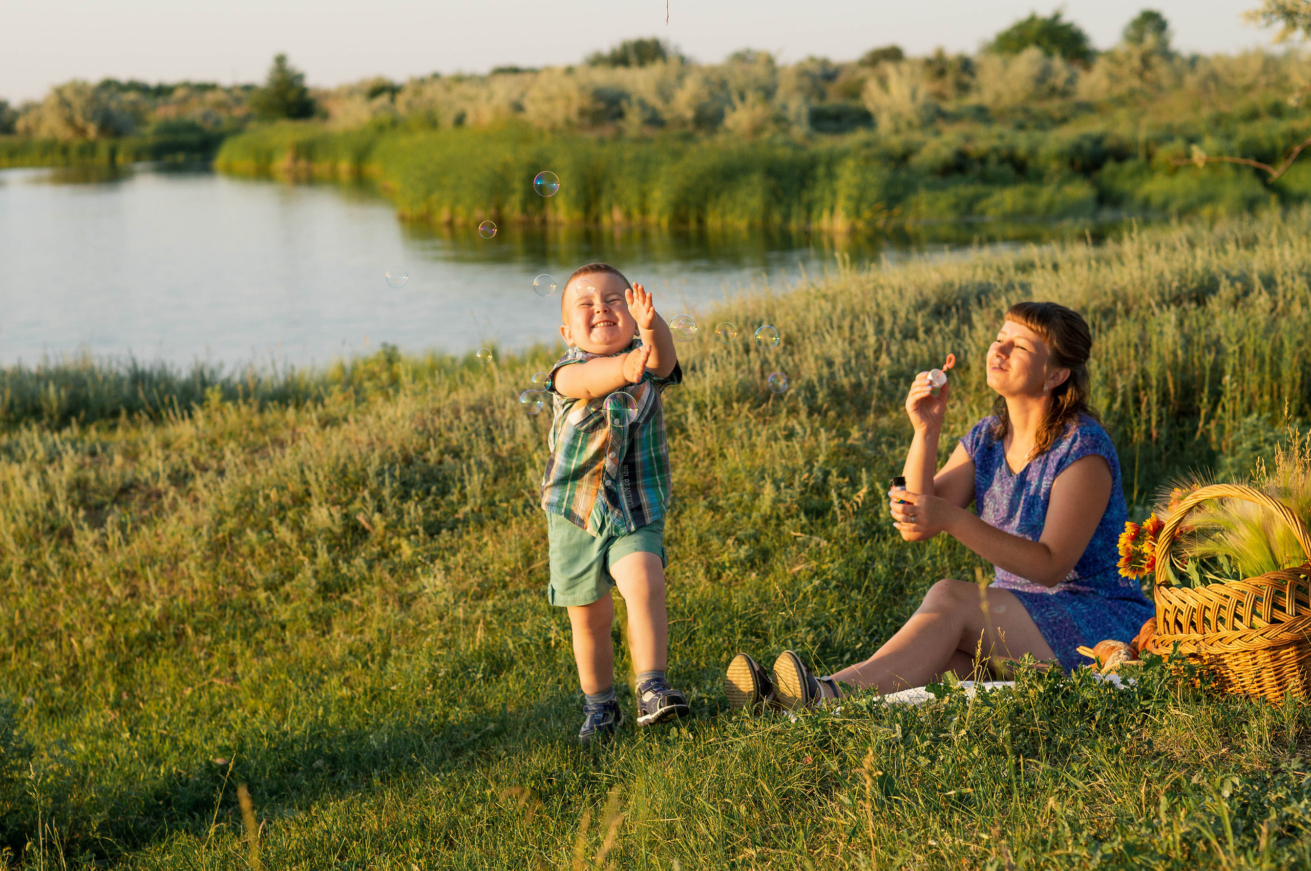 На фоне реки мать пускает мыльные пузыри, а маленький сын очень радуется этому.