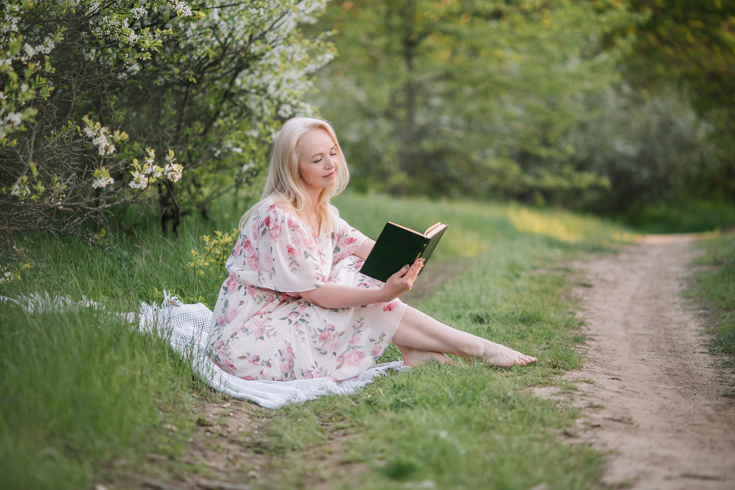 Женщина в белом воздушном платье читает книжку около лесной тропинки.