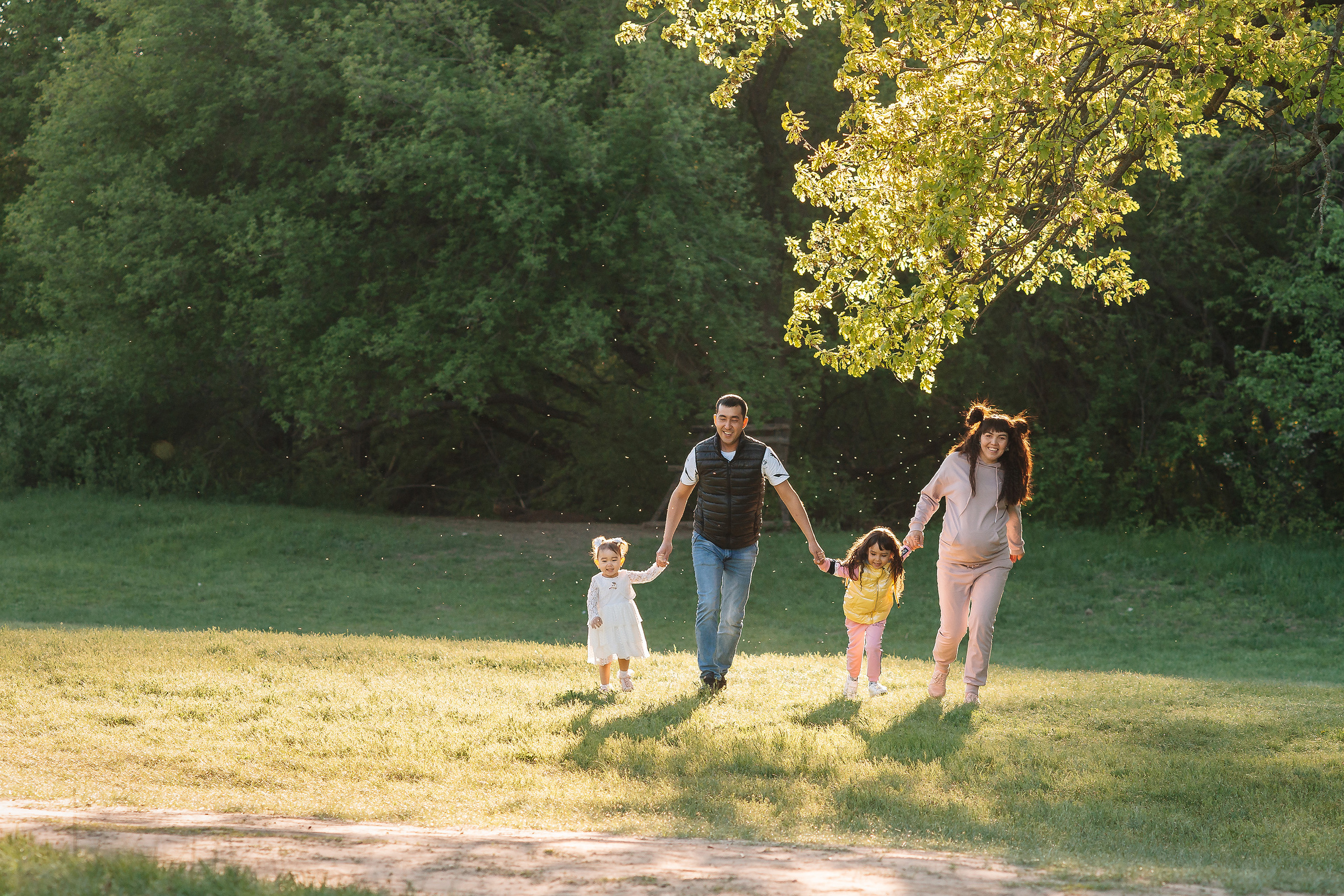 Фотосъемка в Энгельсе прекрасной пары с двумя детьми, бегущей по зеленой лужайки, и освещаемые сзади закатными лучами солнца, а на заднем плане зеленые деревья.