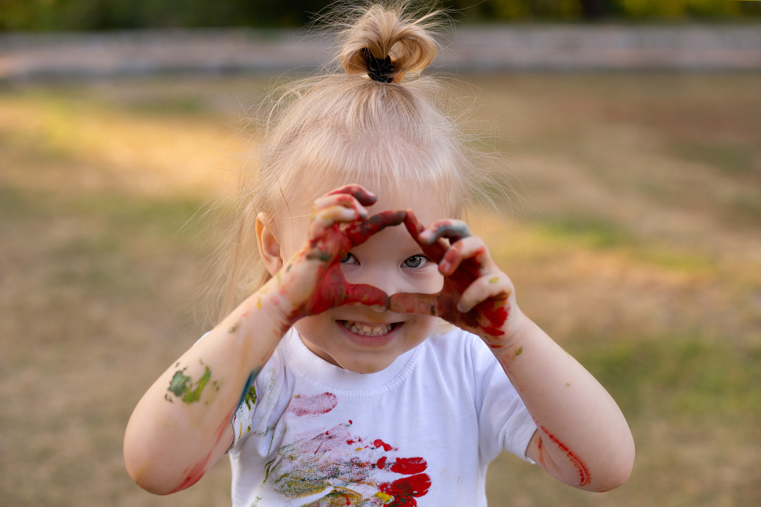 Маленькая девочка в белой футболке вся измазанная краской сложила из пальцев сердечко, и задорно смотрит в камеру.