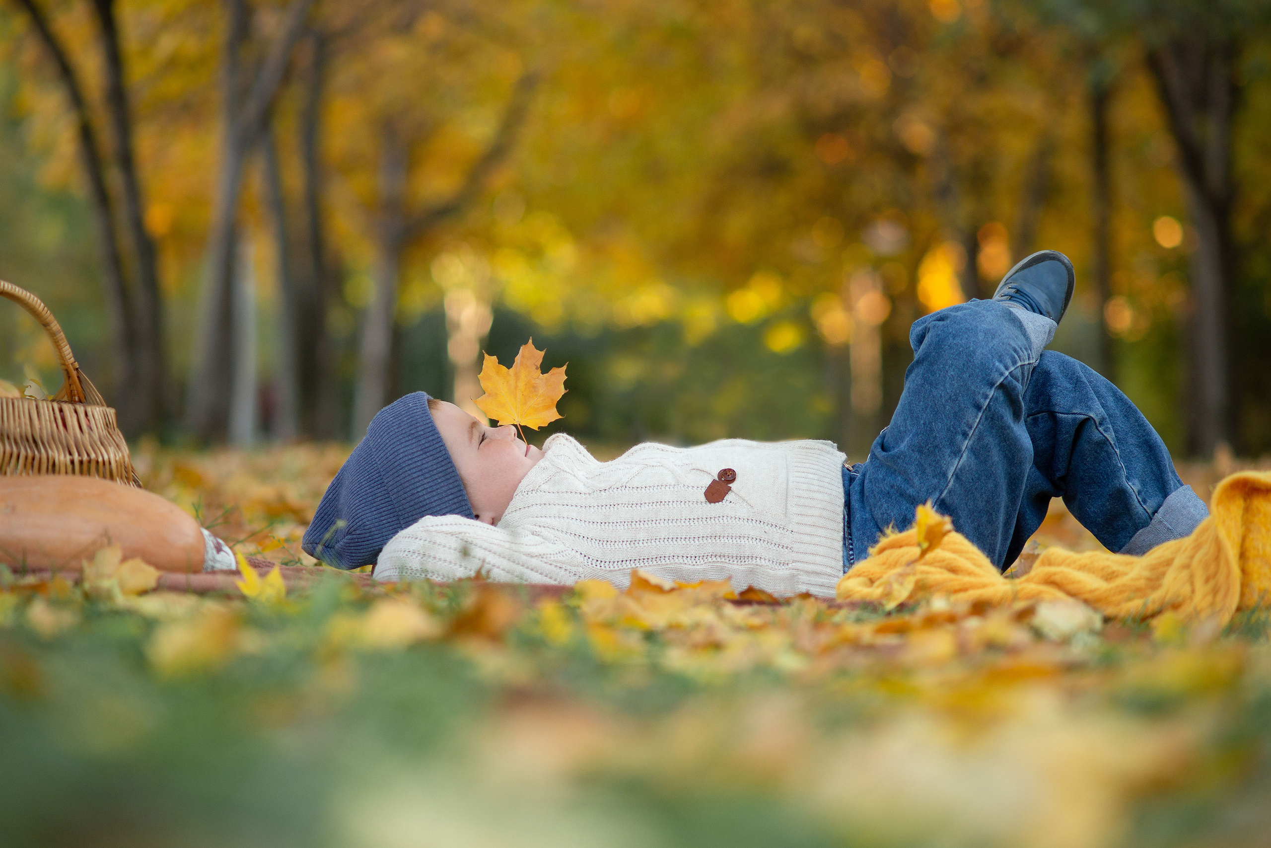 маленький мальчик в белом свитере лежит на желтых листьях запрокинув нога на ногу, и держит в зубах кленовый лист смотря при этом в высь.
