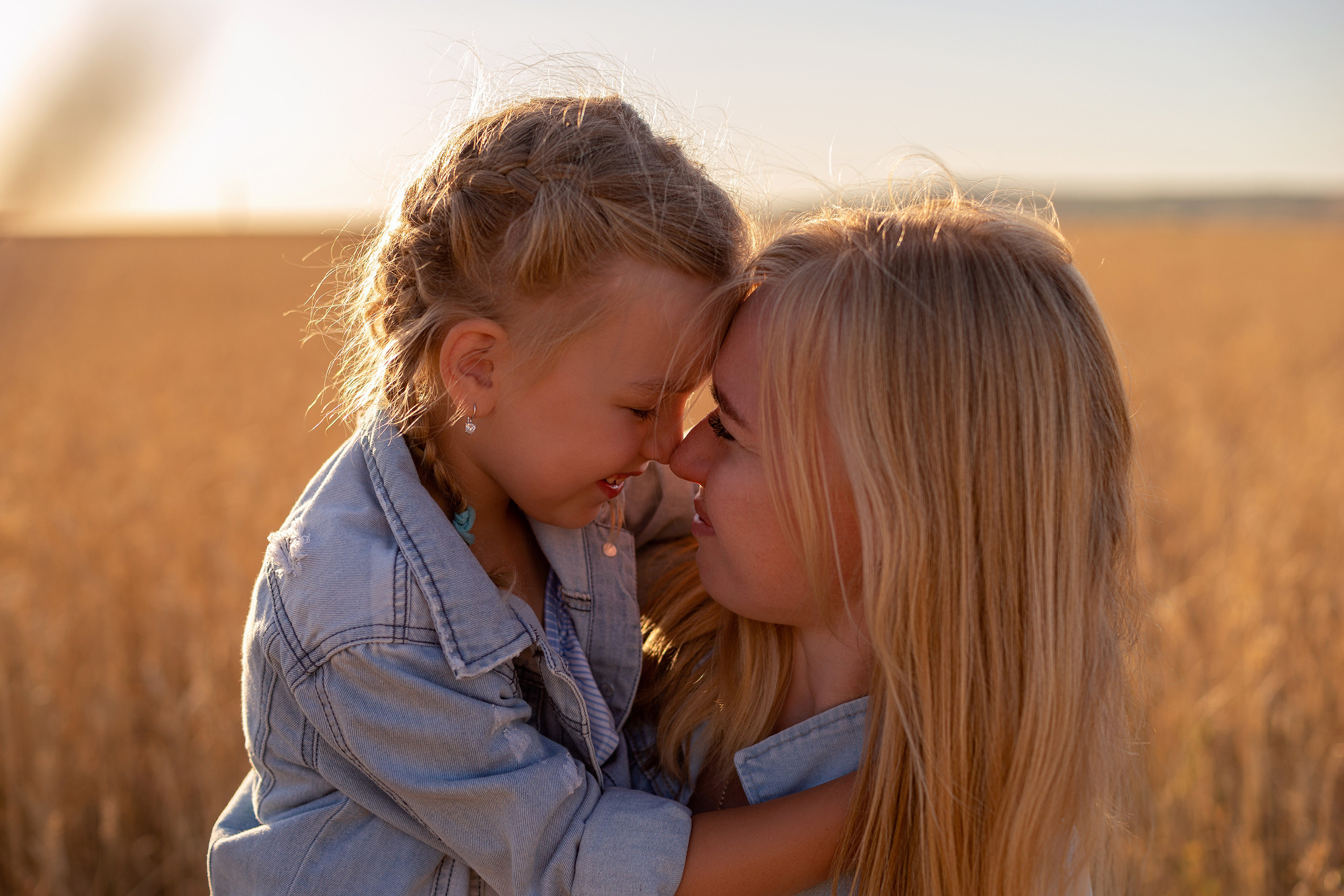 На фото крупным планом девочка обнимает за шею свою маму на фоне пшеничного поля.