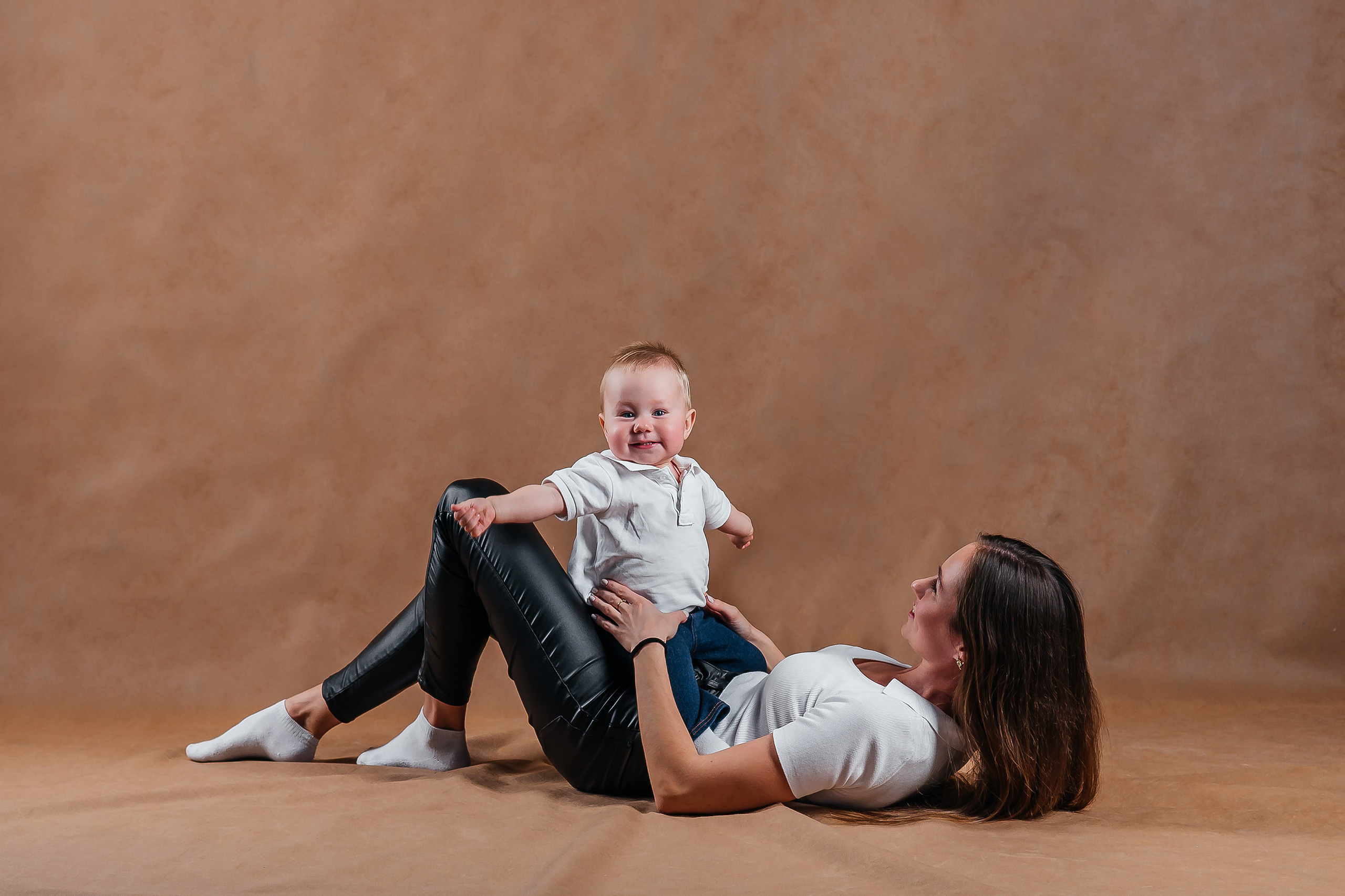 Маленьких мальчик сидя на лежащей матери раскинул руки смотрит в камеру.