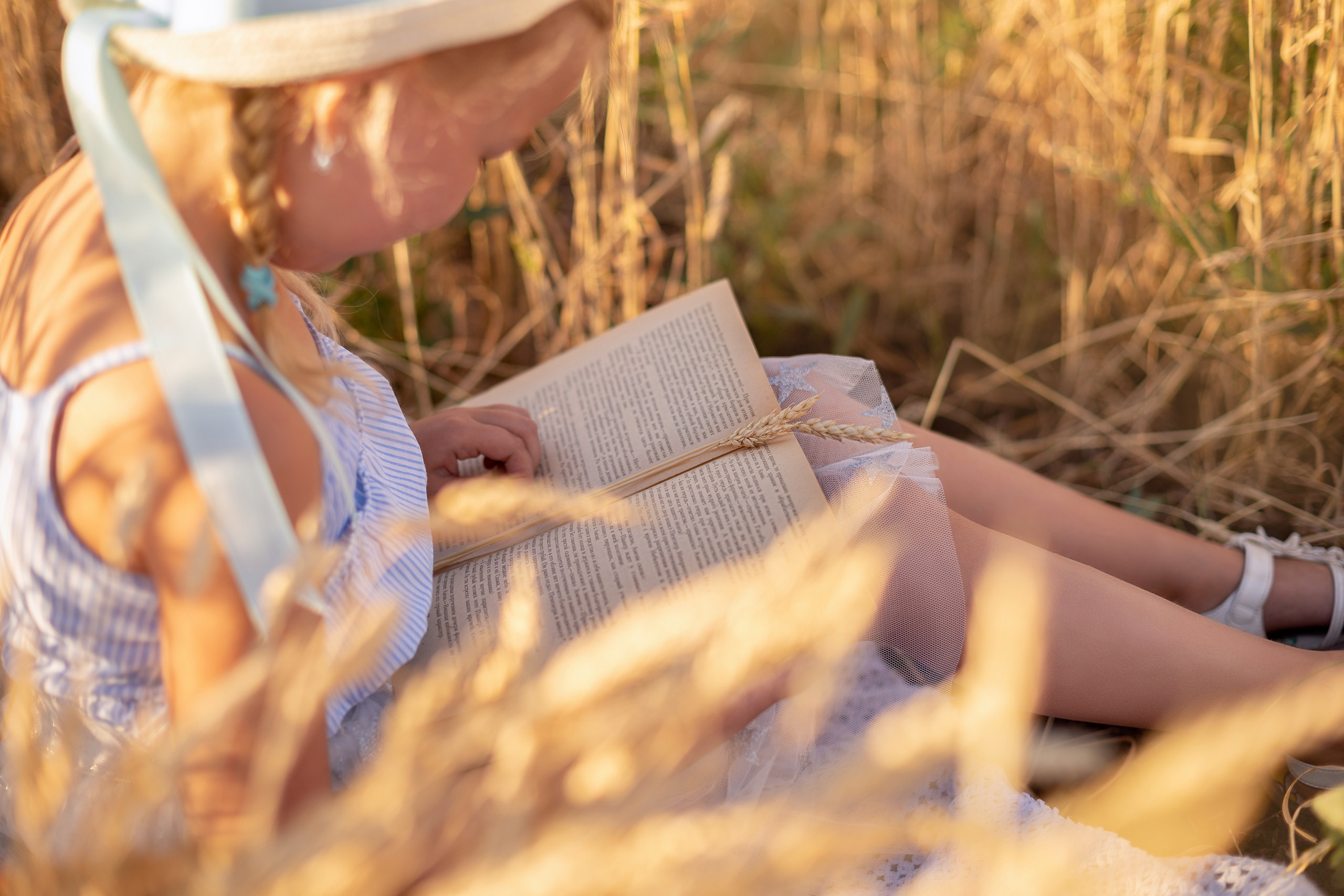 Маленькая девочка в шляпке читает книжку присев посередине пшеничного поля.
