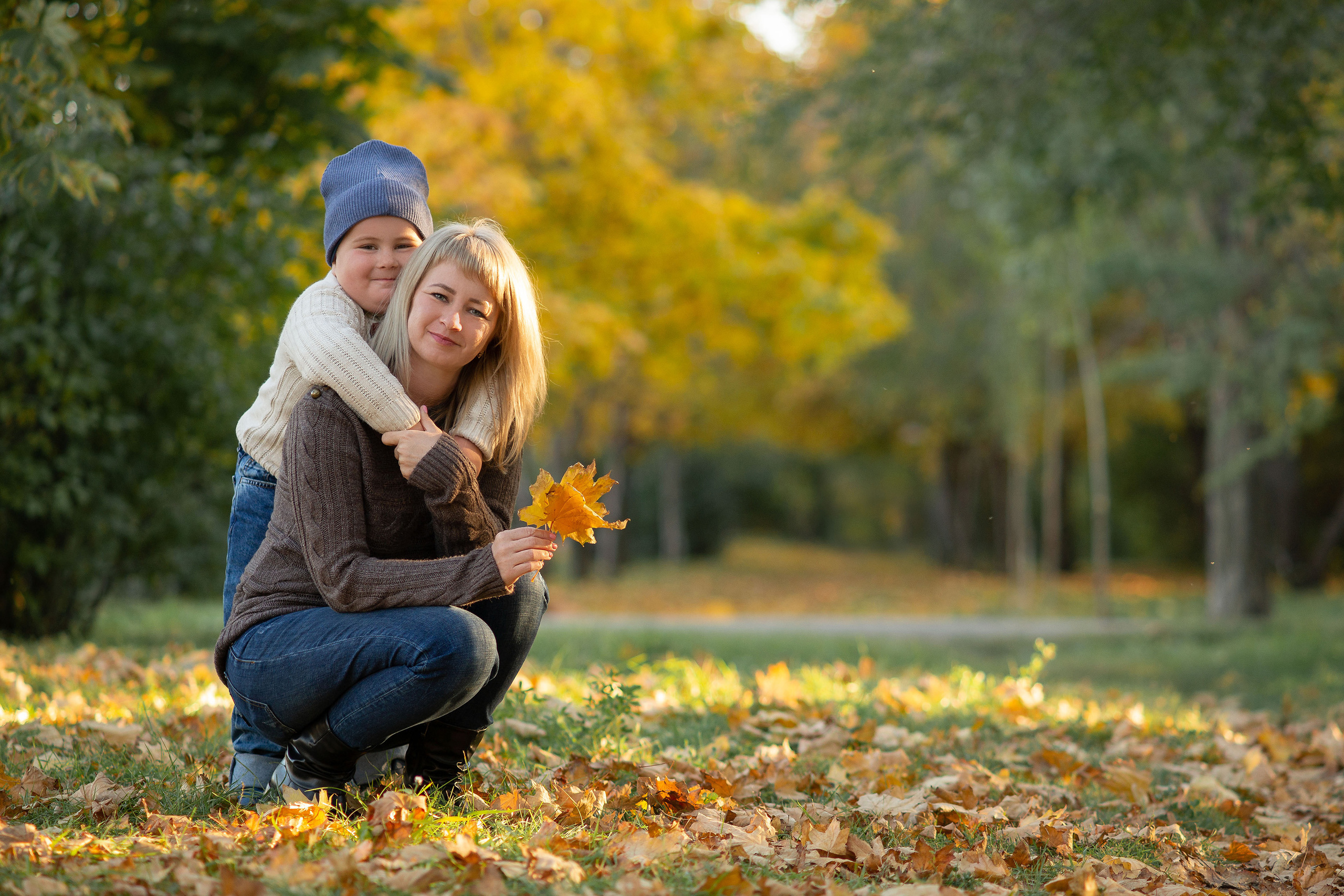 Мальчик мило обнимает сзади маму и они счастливые смотрят в камеру на фоне осеннего леса.