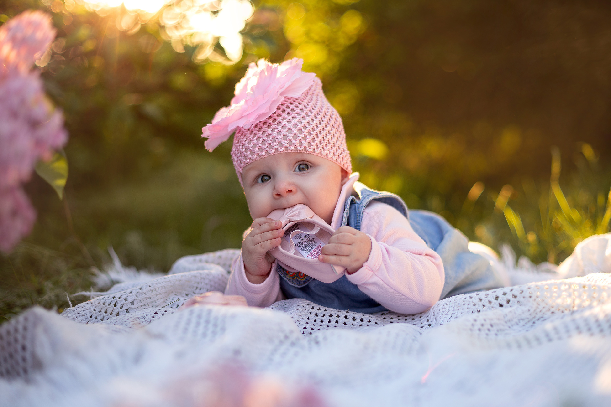 Детский фотограф в Энгельсе сняла маленькую малышку, которая тянет в рот свой башмачок на фоне цветущей сирени.