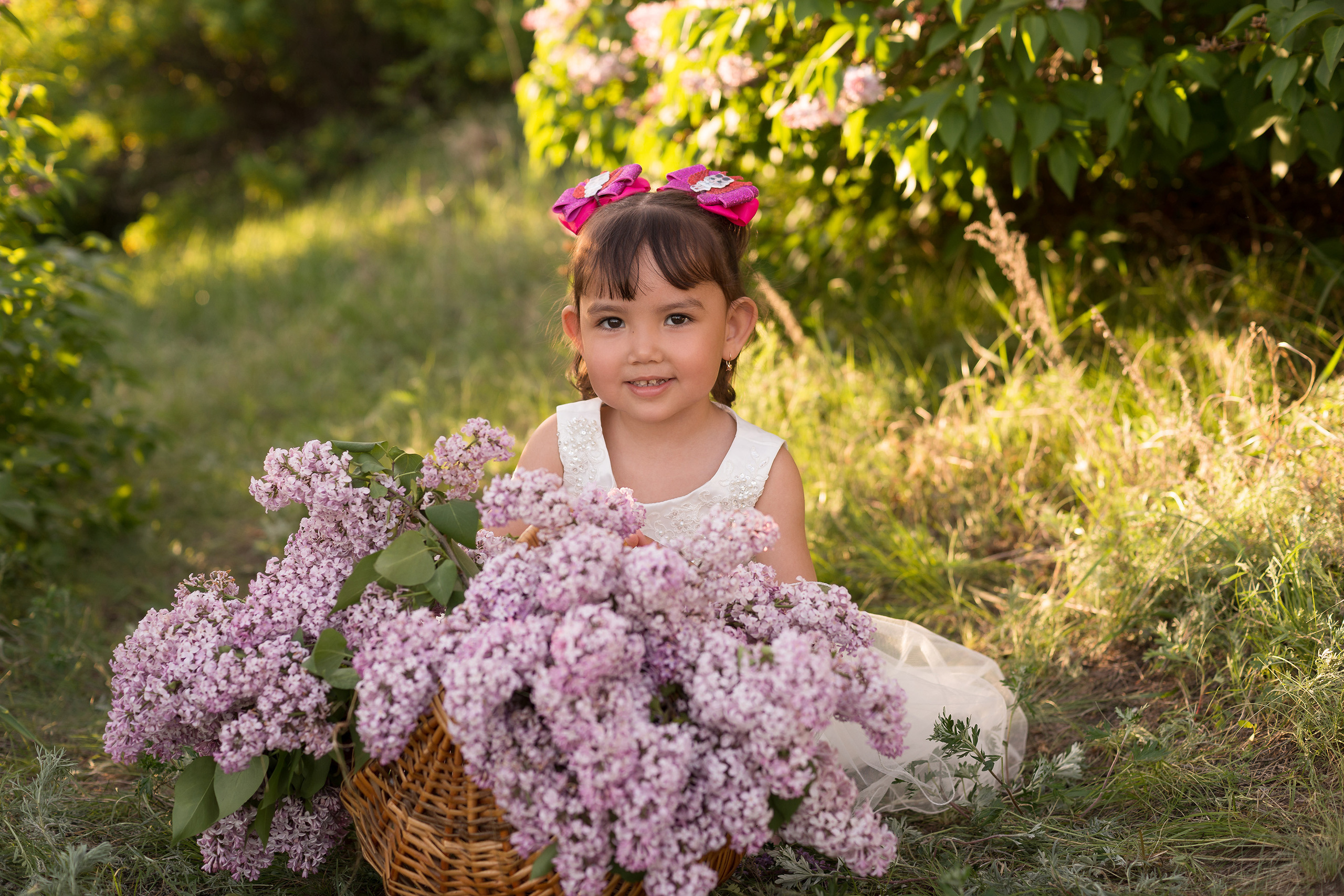 Красивая азиатская девочка в лучах заката сидит рядом с корзиной сирени и улыбается в камеру.