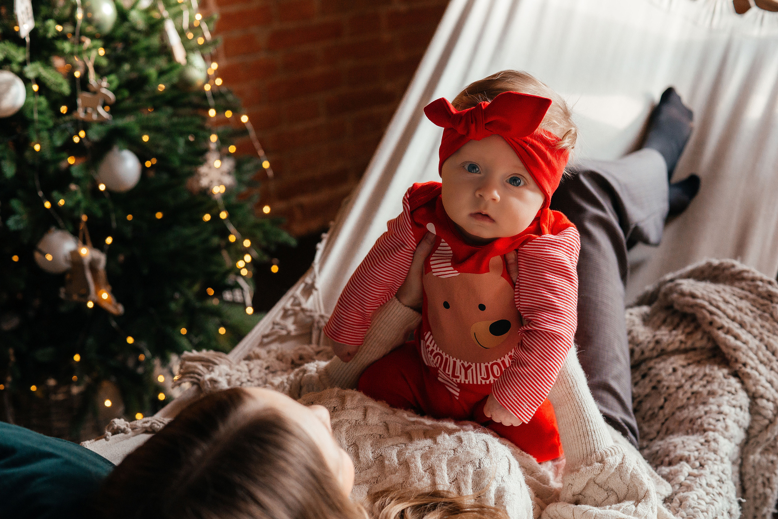 Маленькая малышка  в новогоднем костюмчике сидит на коленях матери в гамаке, с левой стороны новогодняя елка.