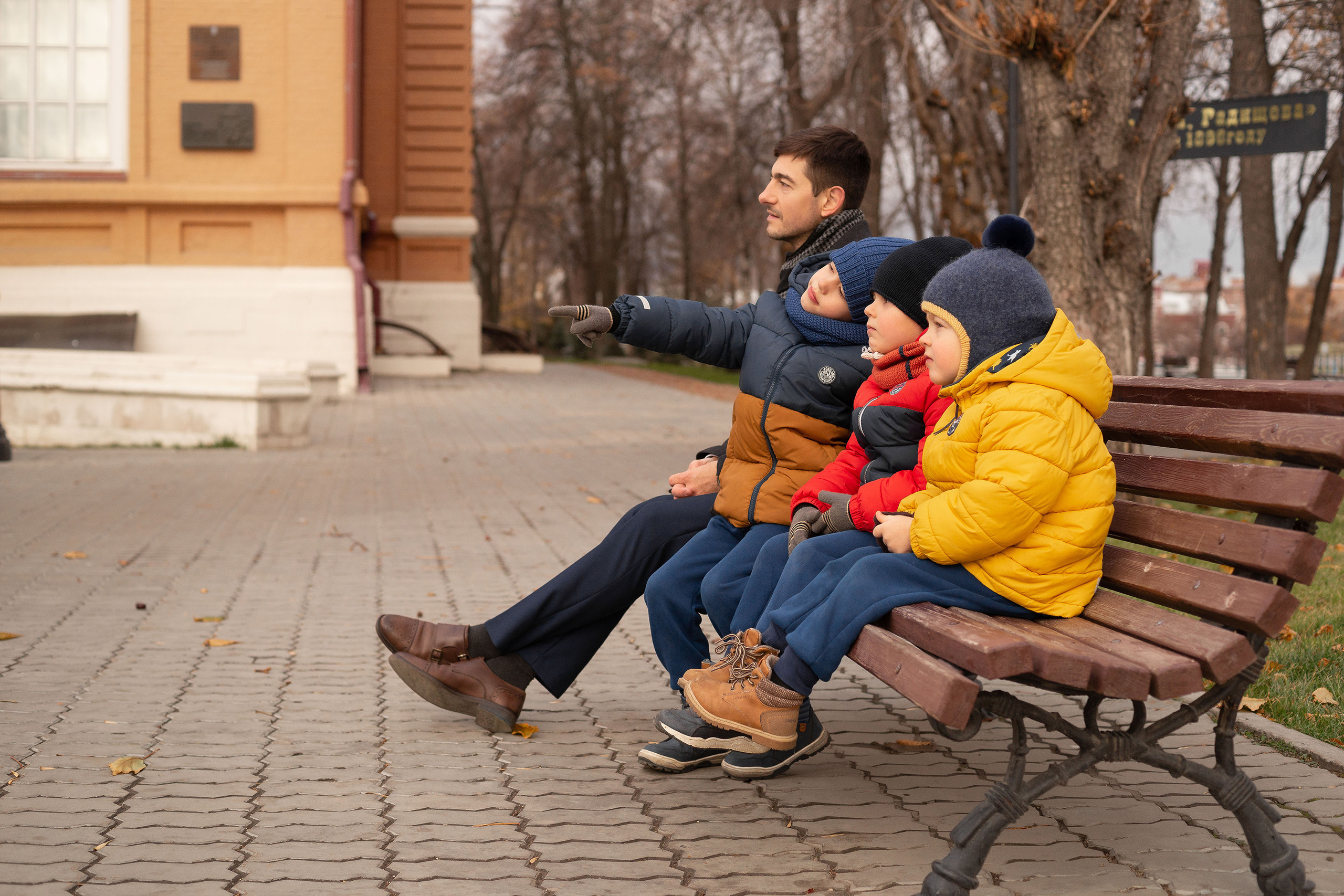 Молодой отец с тремя сыновьями сидят на лавочке в осеннем парке и один из сыновей указывает куда-то вдаль.